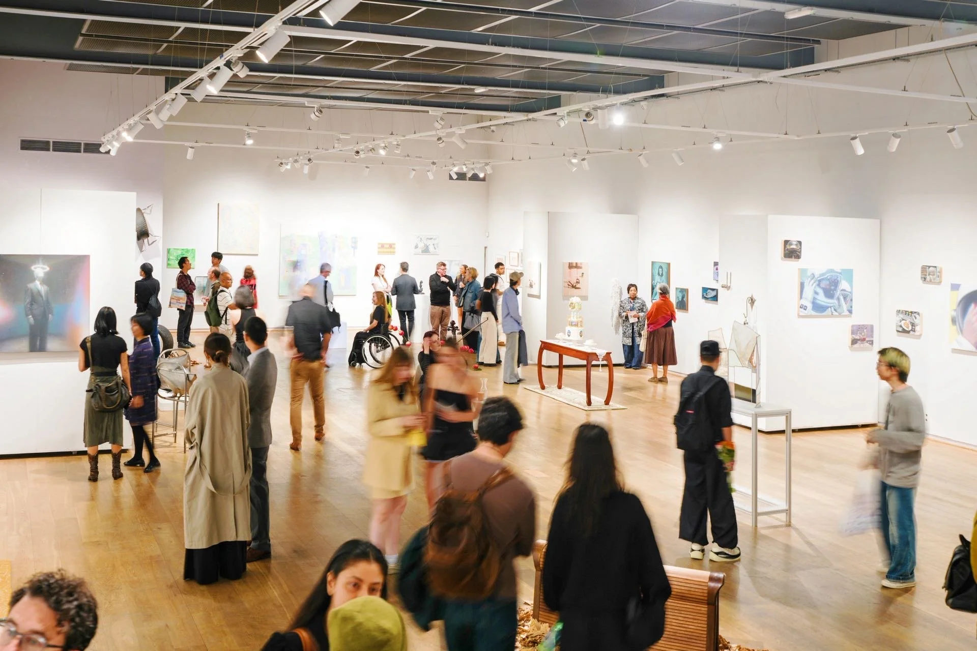 People viewing artwork in a modern art gallery with white walls and wooden floors, informational placards and various art pieces displayed.