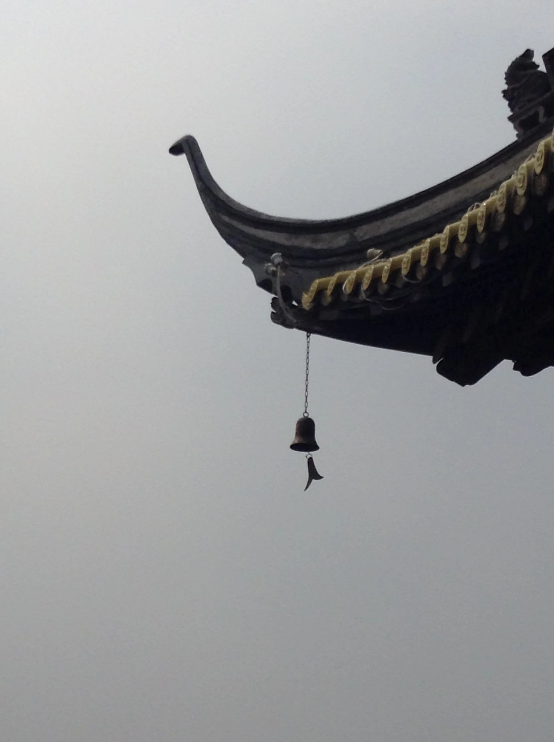 The corner of a traditional Asian style roof with decorative elements and a small bell hanging from a chain, set against a cloudy sky.