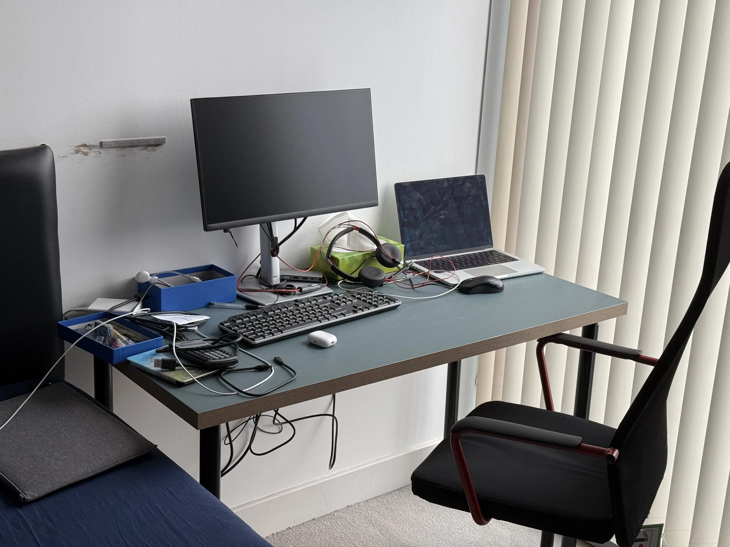 A cluttered office desk with a large computer monitor, a laptop, a keyboard, a mouse, a pair of headphones, and various other electronic accessories and office supplies. The desk is positioned next to vertical blinds covering a window.