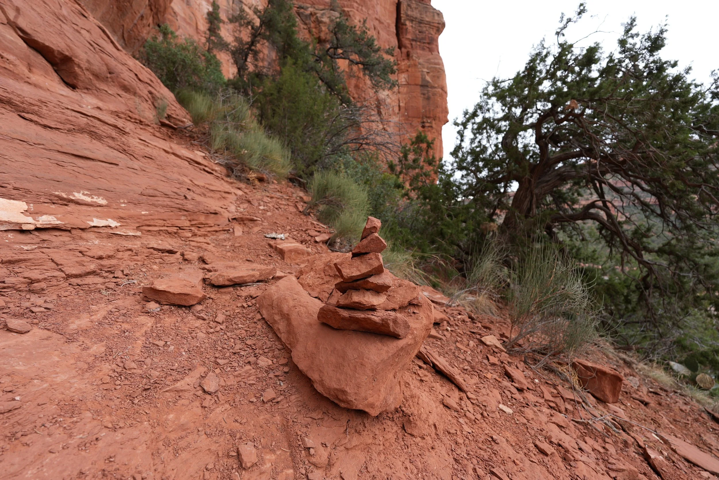 Red rock canyon landscape with stacked rocks, sparse green vegetation, and large trees against a steep, reddish cliff face under an overcast sky.