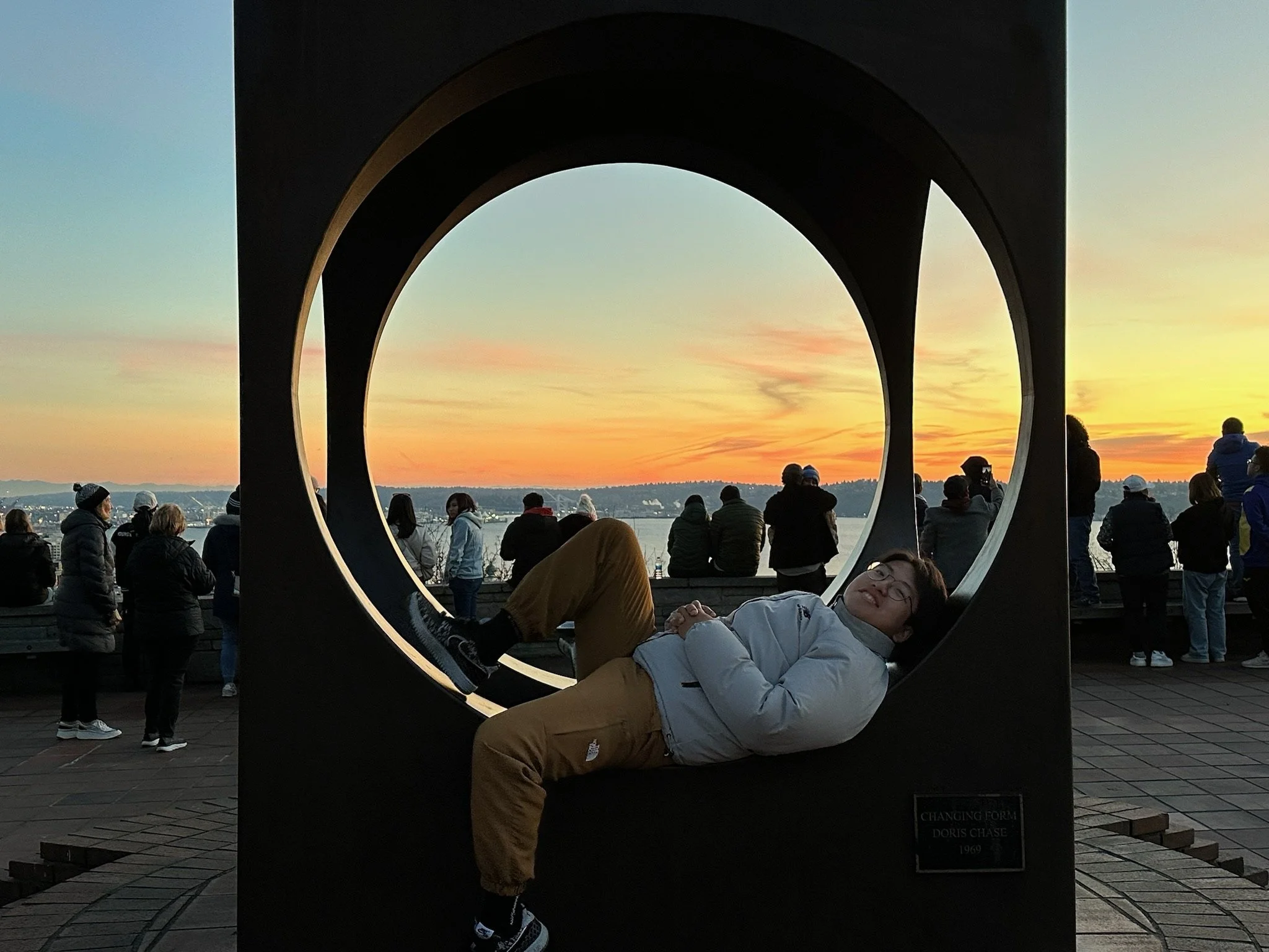A person in a white jacket lying on the ground and resting their head on a large, circular sculpture with a sunset in the background. Several people are standing and looking towards the horizon, some wearing jackets and hats, near a body of water and