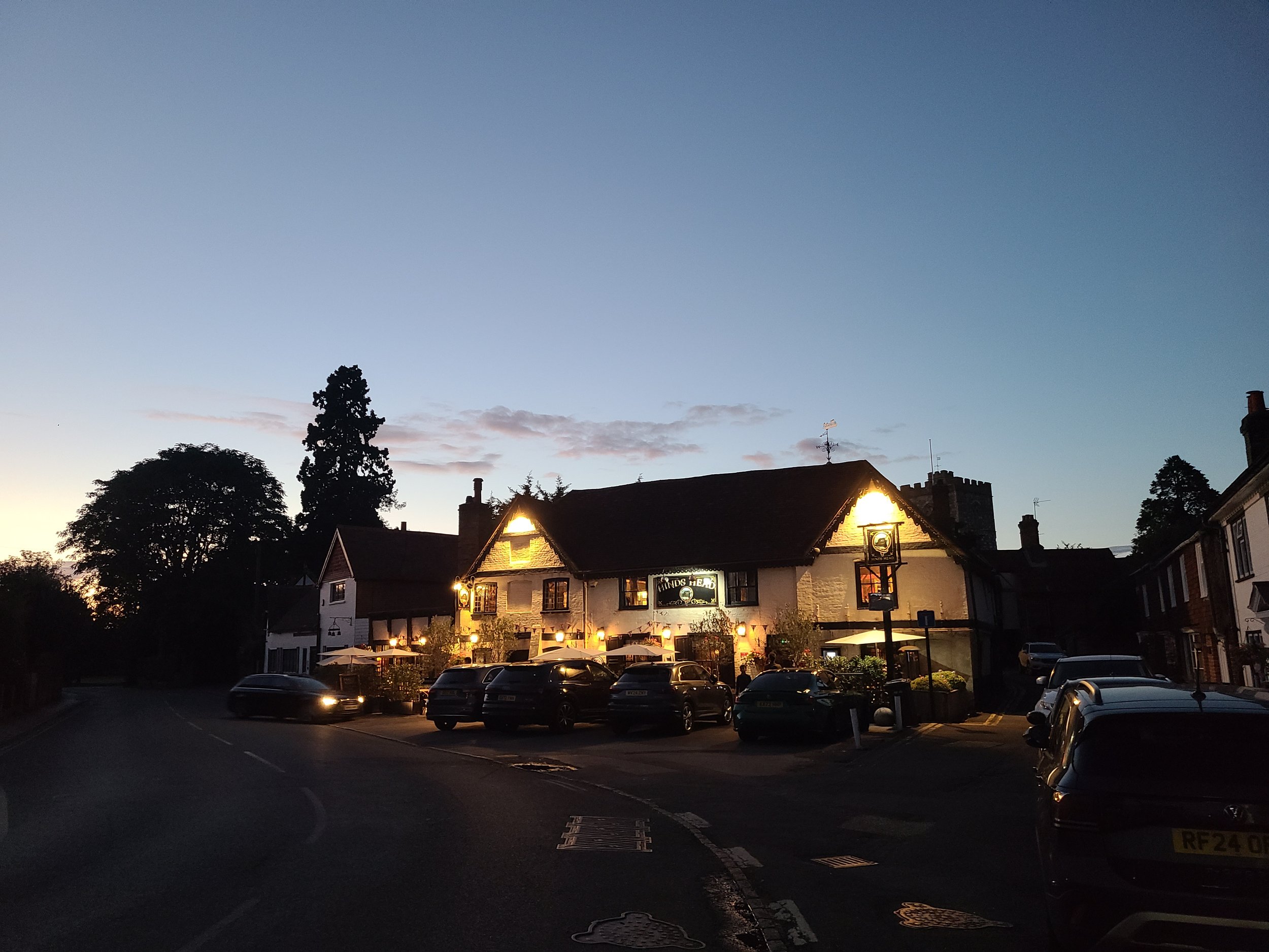 A street view at dusk with parked cars, trees, and a building with lit windows and signage. The sky is transitioning from daylight to night.