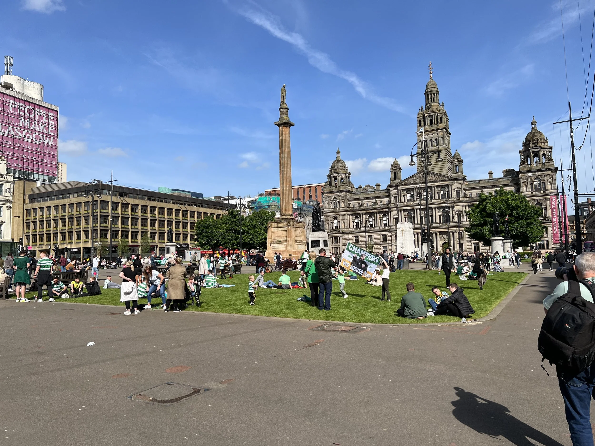 A busy public square filled with people sitting on the grass and walking. In the background, historic buildings and a tall monument with a statue on top are visible under a blue sky with some clouds.