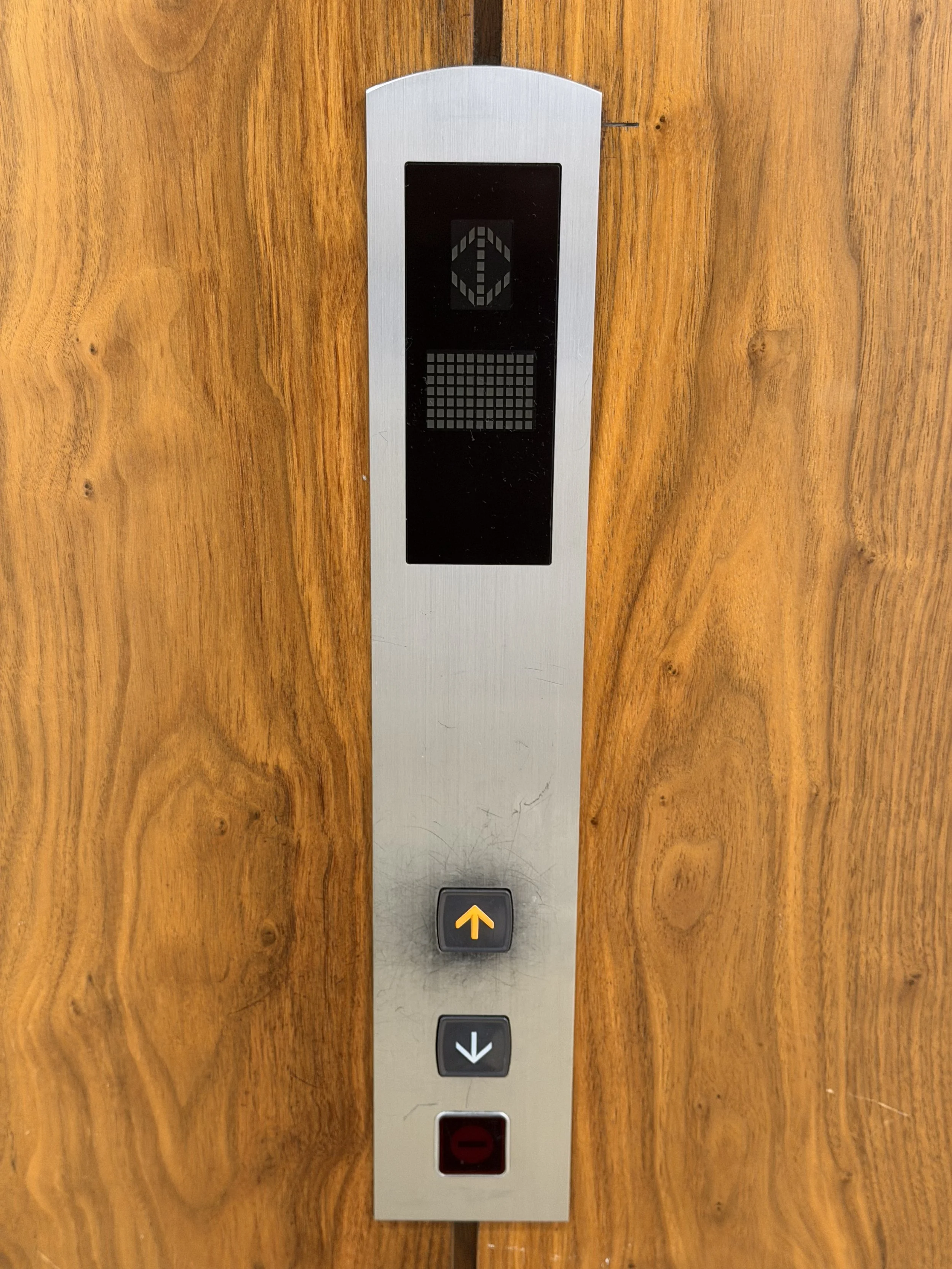Elevator control panel with up and down buttons, a display, and an emergency stop button, mounted on a wooden wall.