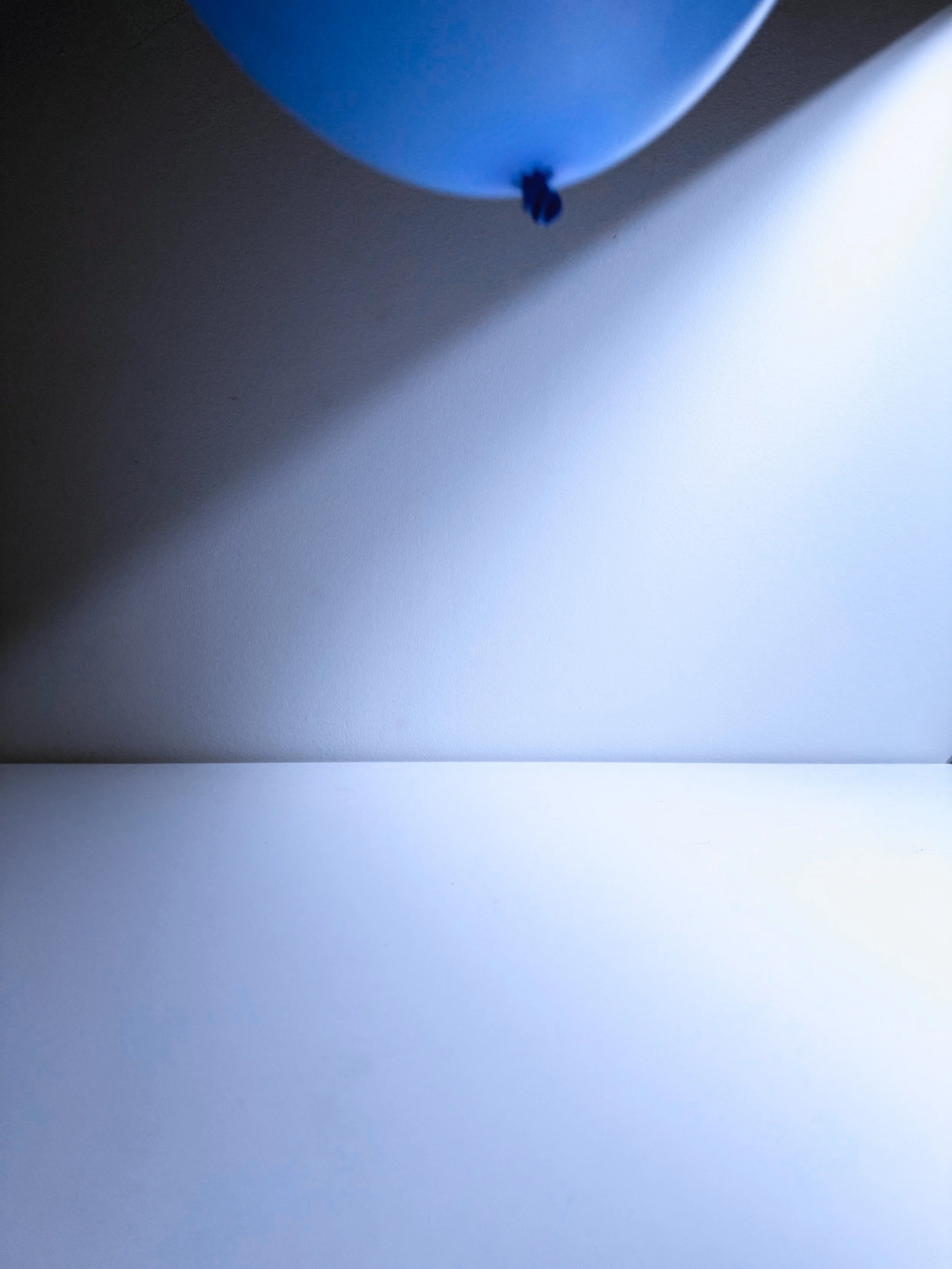Close-up of a blue balloon with a small knot at the bottom, casting a shadow on a plain white wall.
