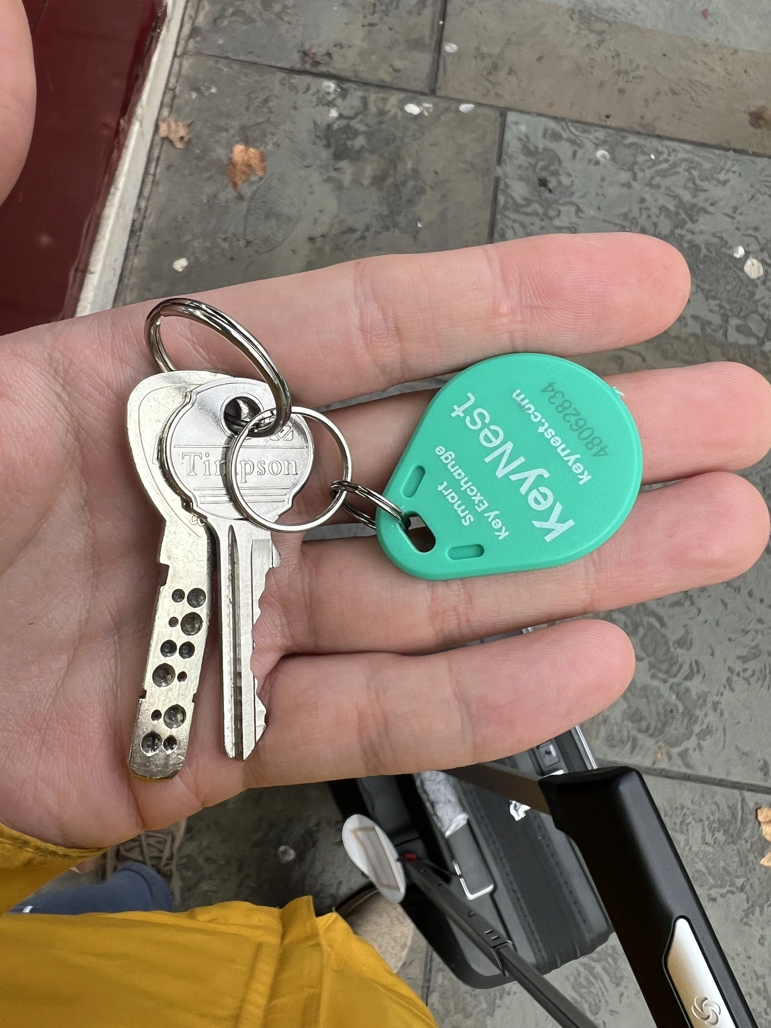 A person's hand holding a set of keys, a green key tag reading 'KEYS' and a key with a unique, dotted pattern is visible. The person is wearing a yellow jacket, and a portion of a black vehicle or luggage is seen in the background on a tiled floor.