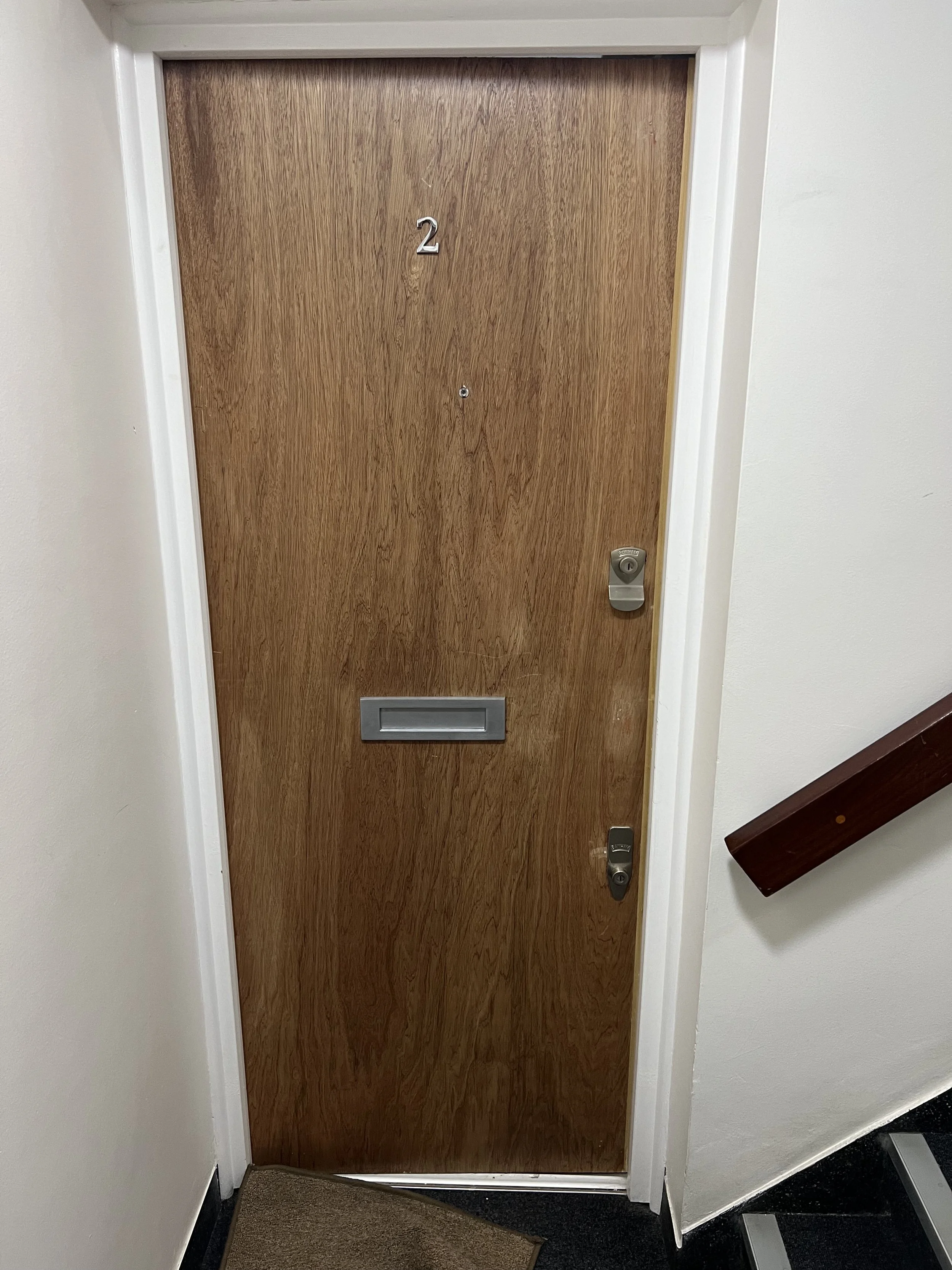 A wooden apartment door with the number 2, a mail slot in the middle, and a silver doorknob and lock on the right side. The door is set in a white wall with a dark wooden handrail on the right side and a small brown and black carpeted step in front.