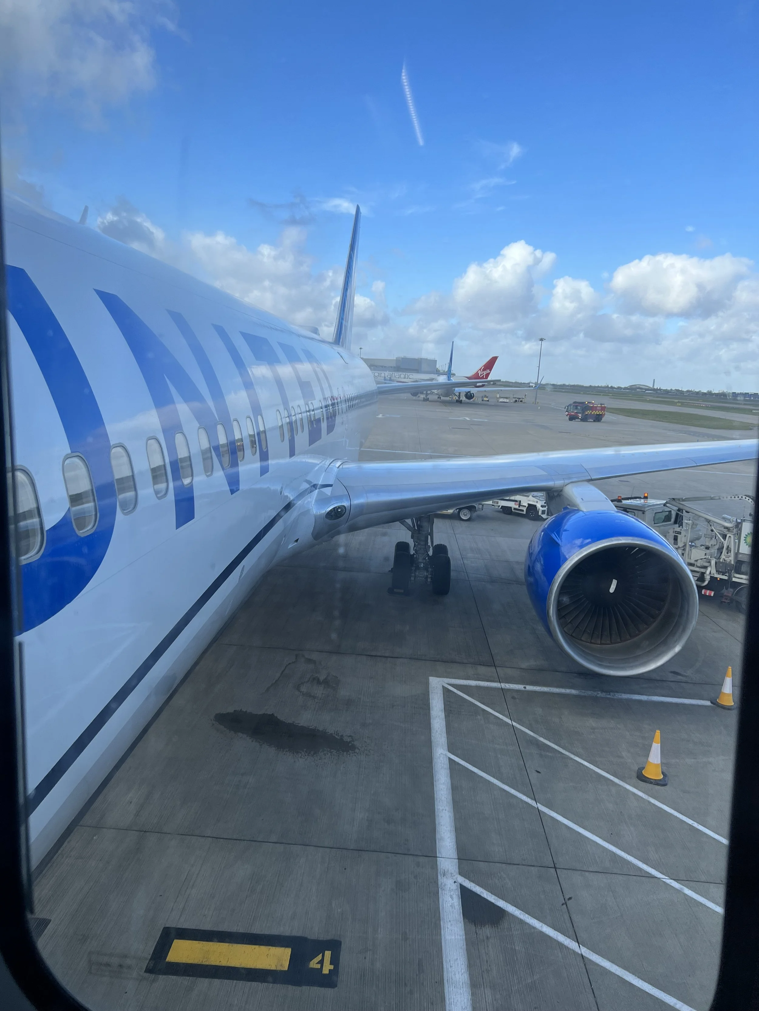 View of airport tarmac with airplane, including blue engine and part of fuselage, and another airplane with Virgin logo in the background. Sky with scattered clouds.