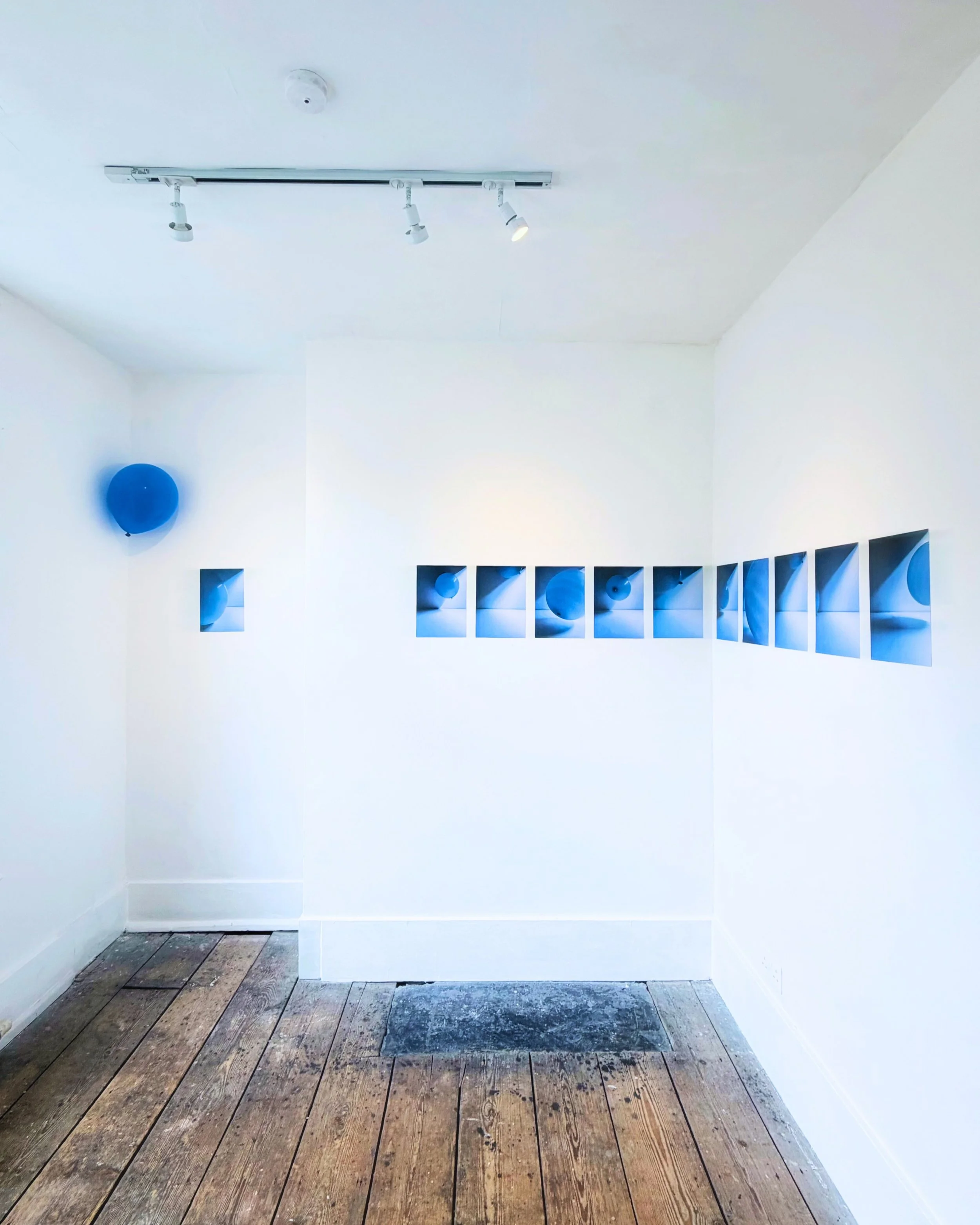 A white gallery space with a row of square display niches on the wall, blue balloons in some niches, a blue balloon on the left wall, and track lighting on the ceiling illuminating the space. The floor is made of aged, weathered wooden planks.