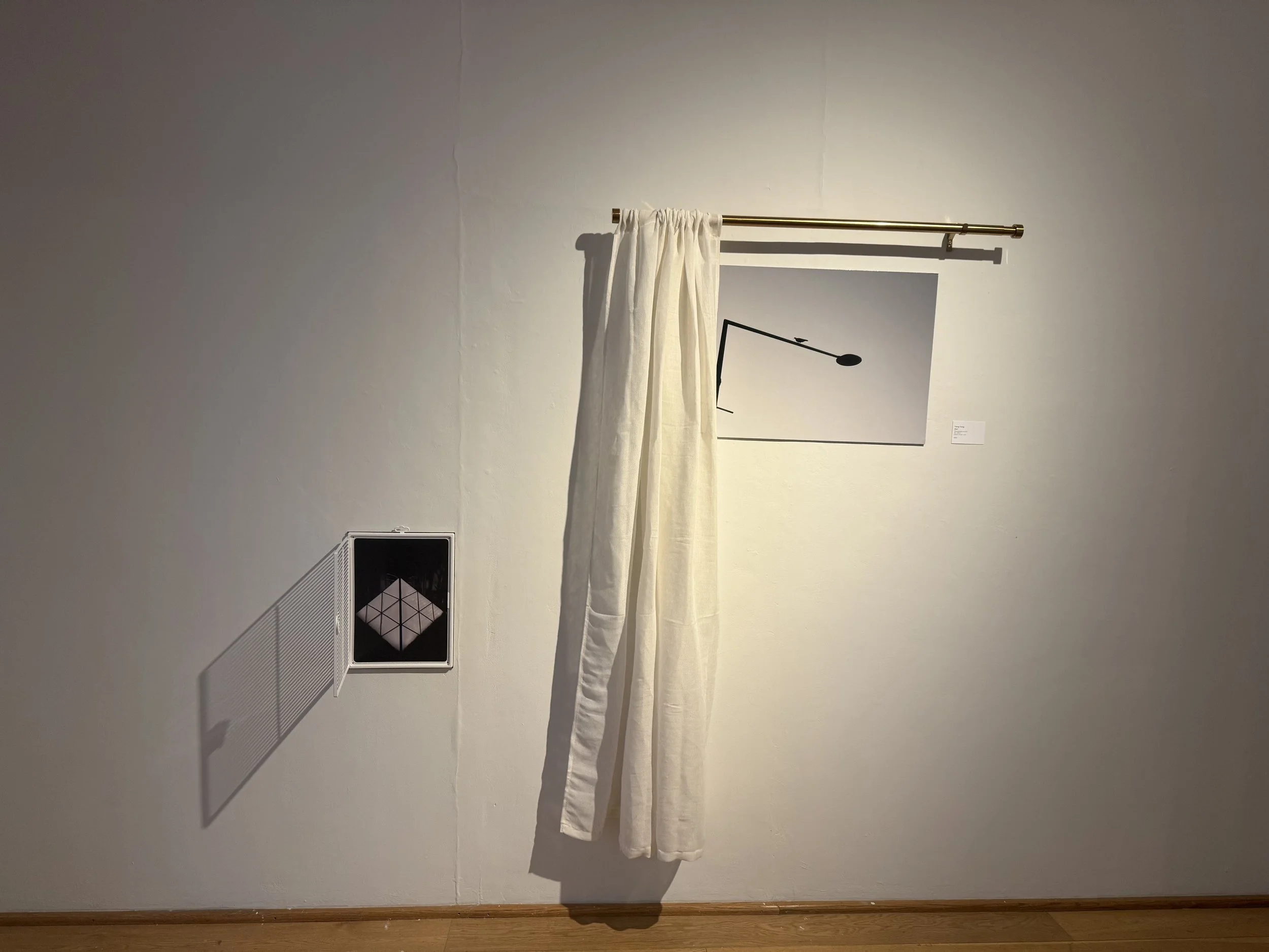 A white curtain hanging from a gold curtain rod in front of a framed black and white photograph on a white wall. There is a vent on the lower left side of the wall and a small nameplate or label next to the photograph.