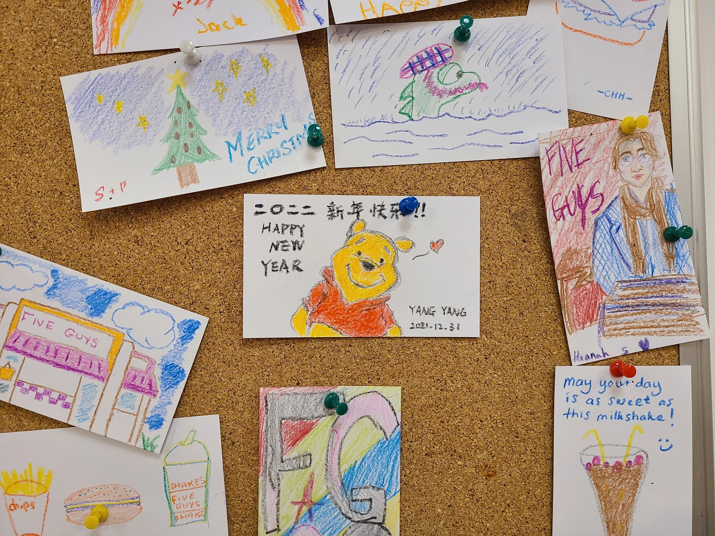 A bulletin board with various children's drawings and handwritten notes, including Christmas trees, a frog on a lily pad, a bear, a person, and messages like 'Merry Christmas,' 'Happy New Year,' and 'Five Guys.'
