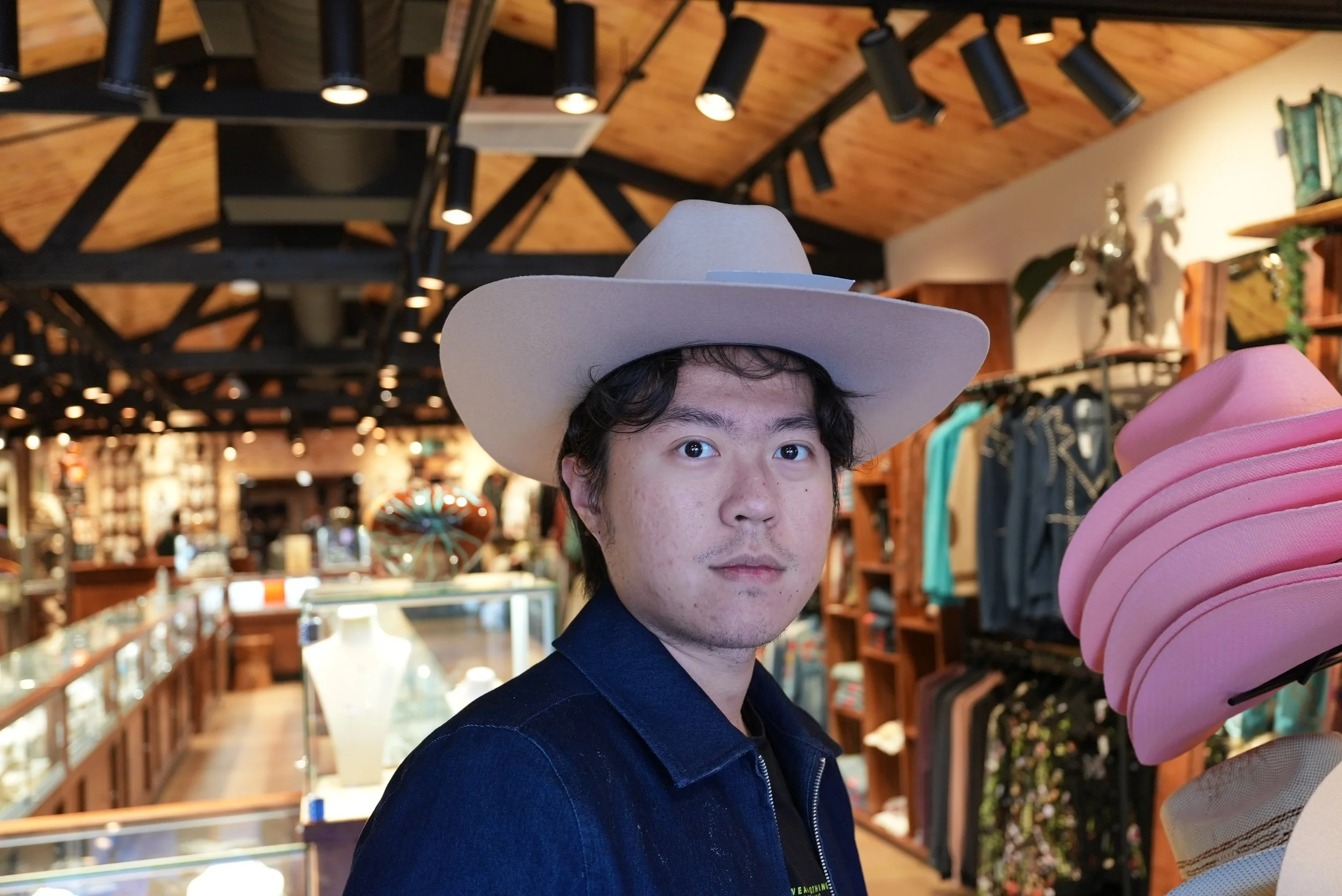 A young man with dark, wavy hair wearing a white cowboy hat and a dark jacket standing inside a clothing store, with store shelves and clothing racks in the background.