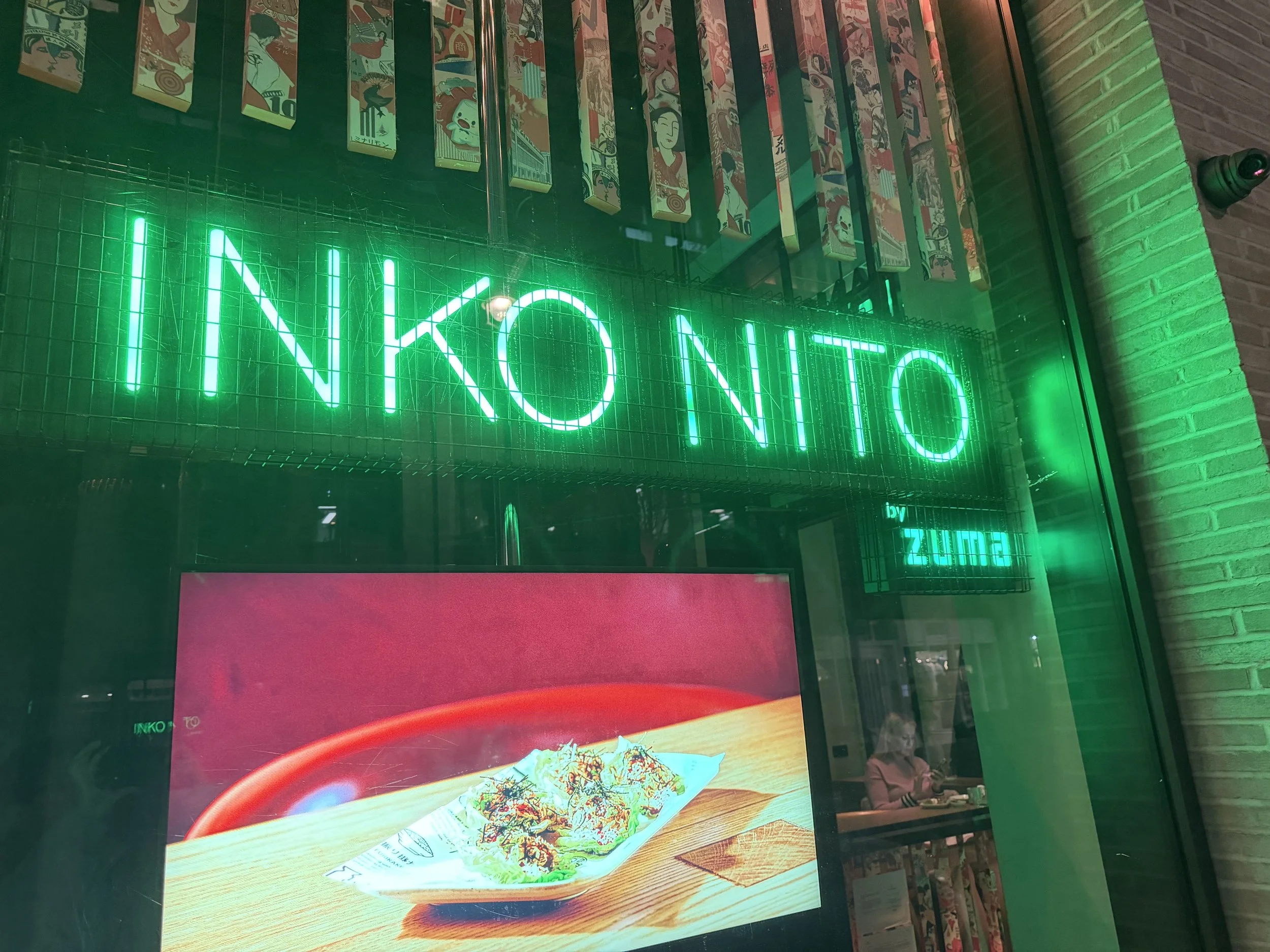A neon green sign spelling 'INCONTO' on a glass window at night. Reflections of a person sitting inside a restaurant can be seen.