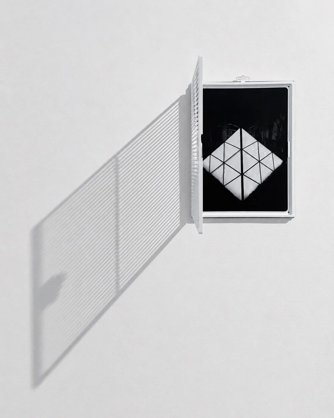 A small, square mirror with a white frame mounted on a plain white wall, reflecting a geometric pattern created by a cube-shaped object and a grid, with a vent or shutter to the side casting a striped shadow.
