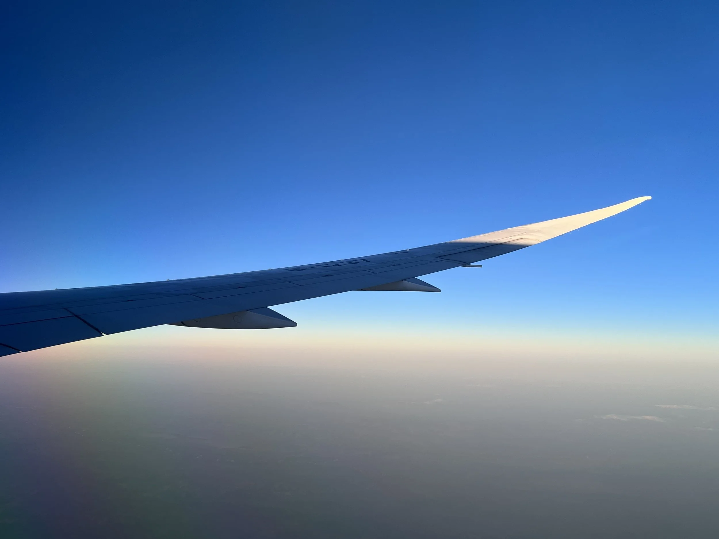 View of an airplane wing flying above clouds with a clear blue sky.