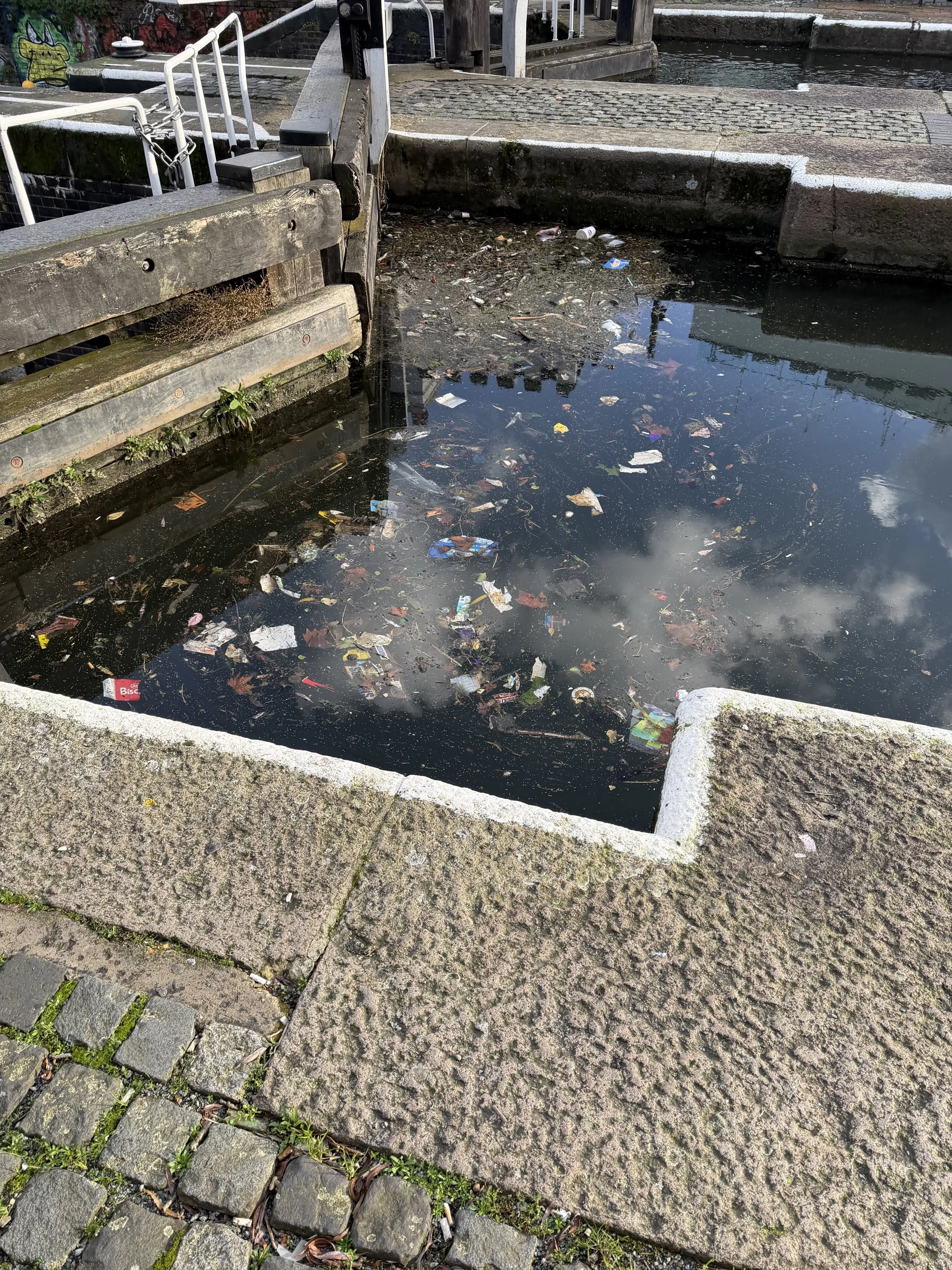 A canal with stagnate water filled with trash and debris, bordered by stone and brick walkways, with a bridge and graffiti in the background.
