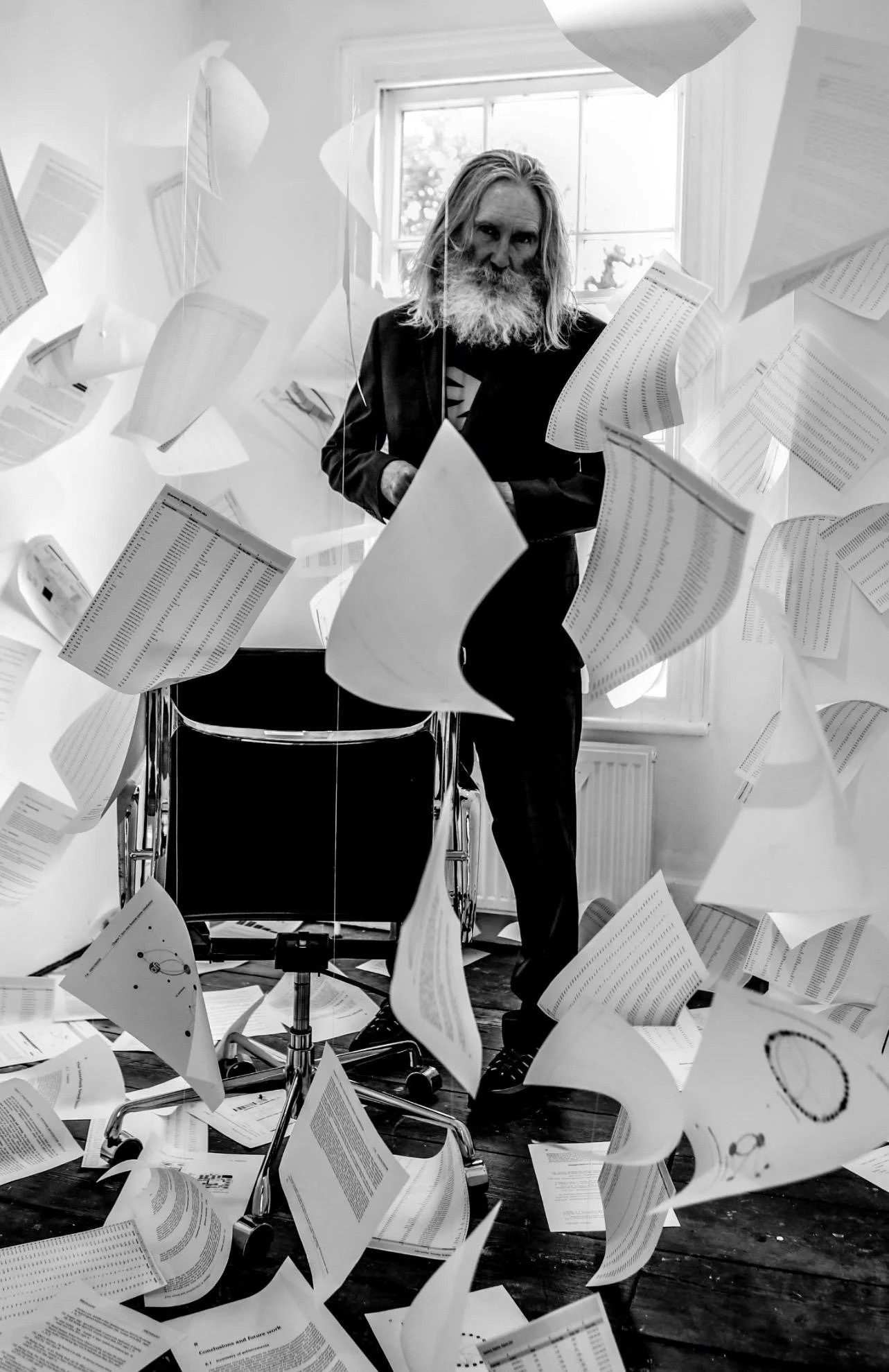 Man with long hair and beard standing in a room with papers flying around him.