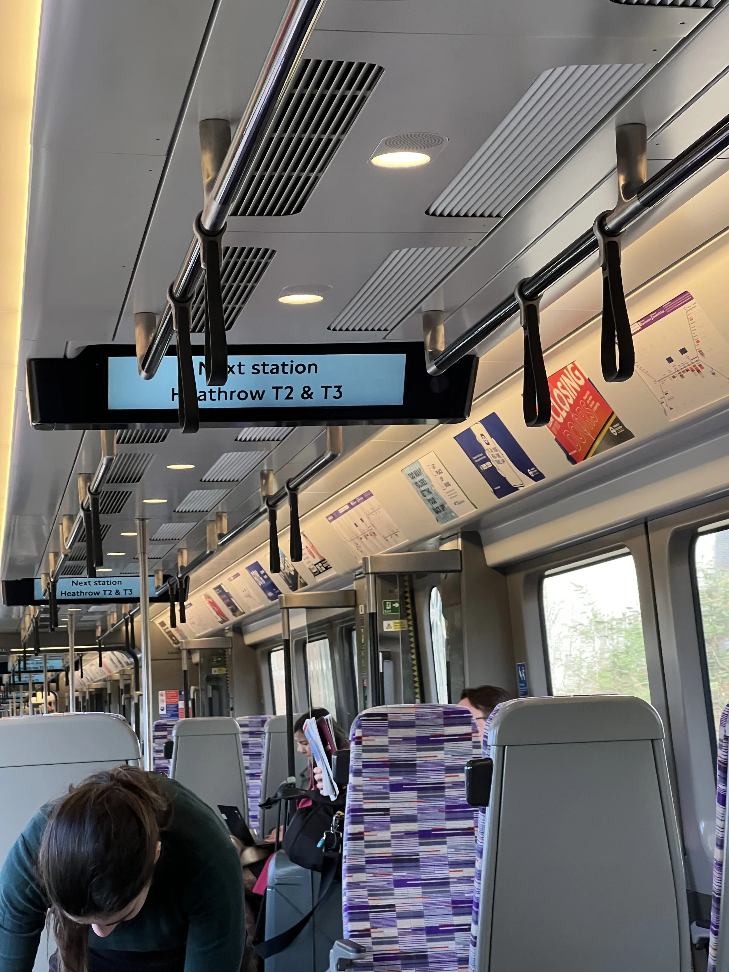 Inside a train, passengers are seated and reading. Overhead signs display next station information for Heathrow T2 & T3. The train has patterned seats, and some passengers are visible, including a woman with dark hair reading a magazine and a person 
