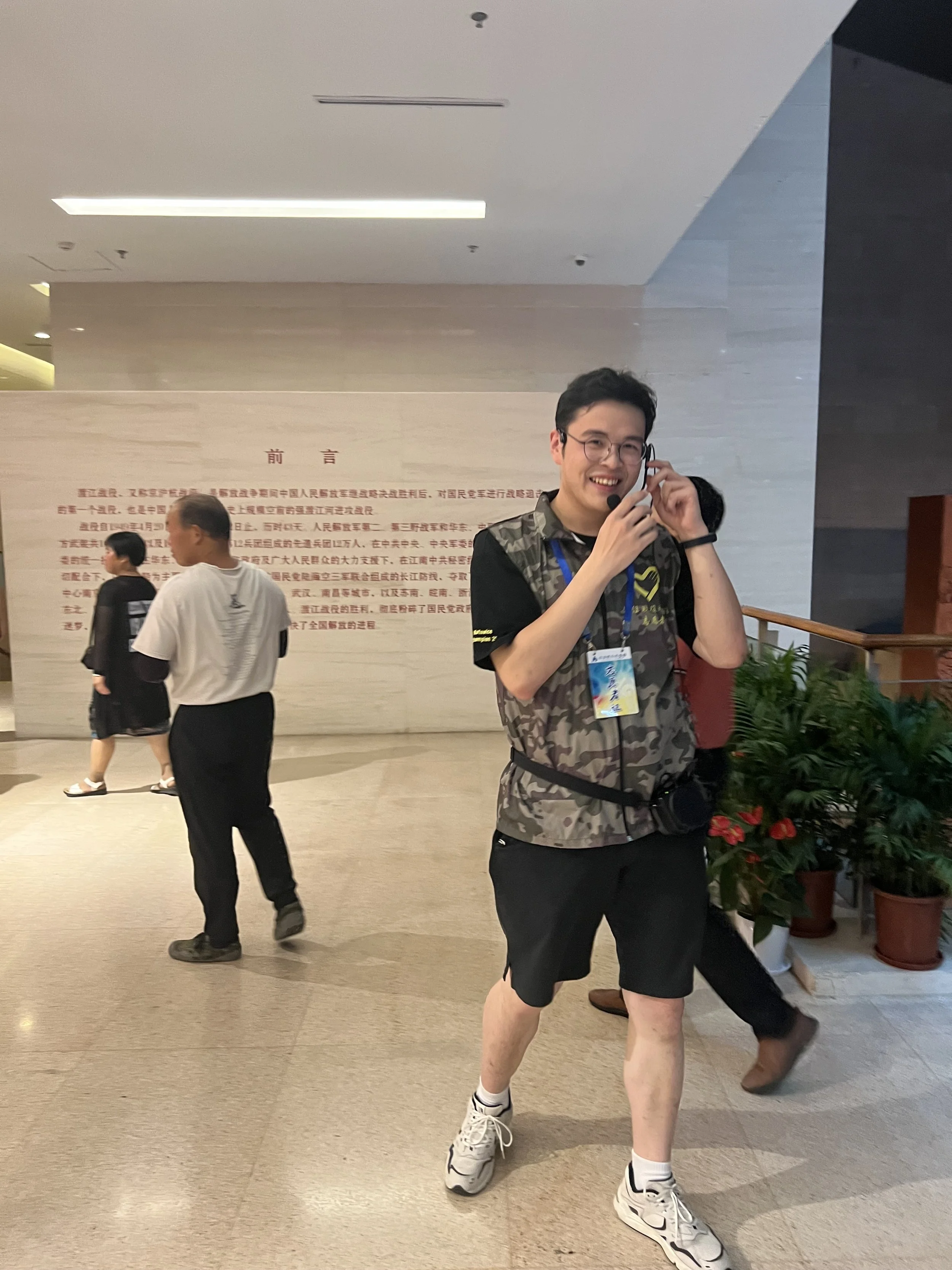 Young man with glasses, wearing a camouflage shirt, black shorts, white sneakers, and a headset microphone, standing indoors near potted plants and a wall with red Chinese characters, smiling and talking.