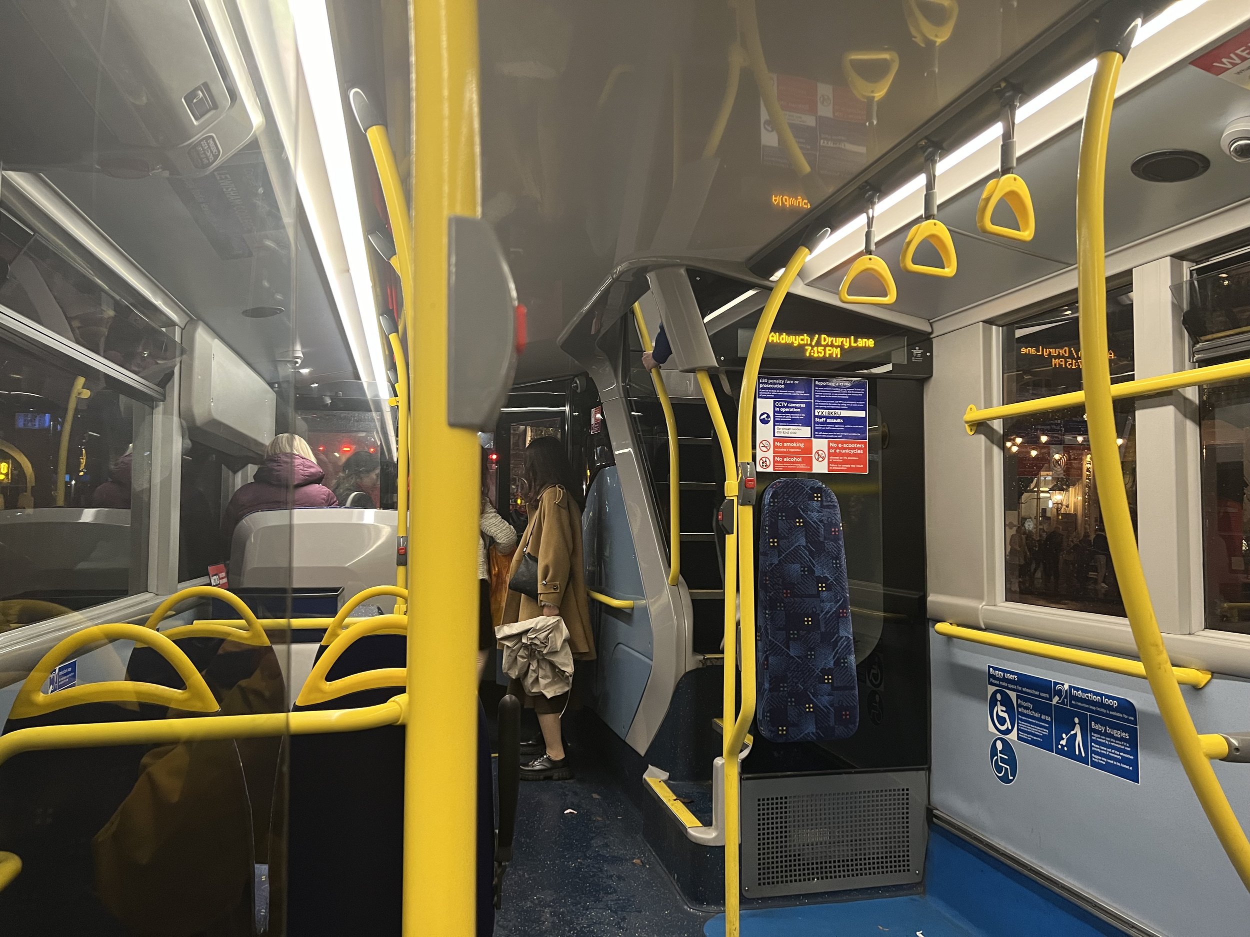 Inside a city bus, showing yellow handrails, passengers standing, and an electronic sign mentioning the bus route and time.