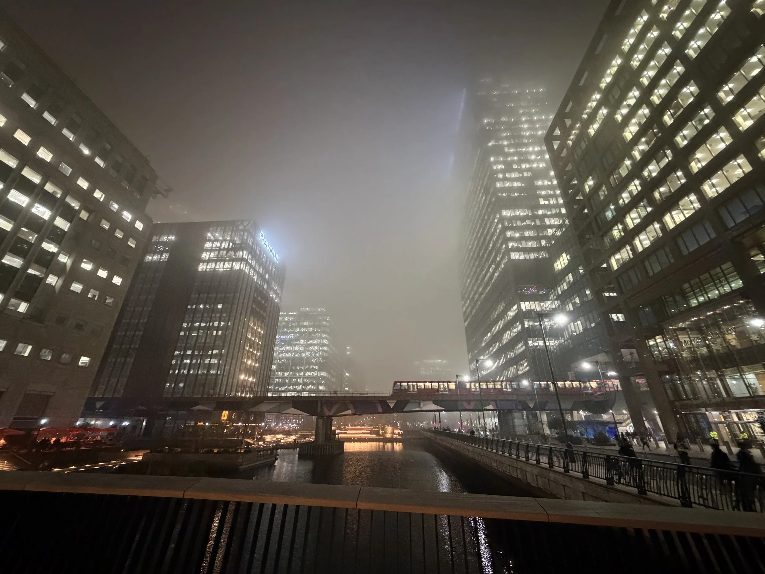 Nighttime city scene with fog, modern high-rise buildings with lit windows, and reflections on the wet pavement.