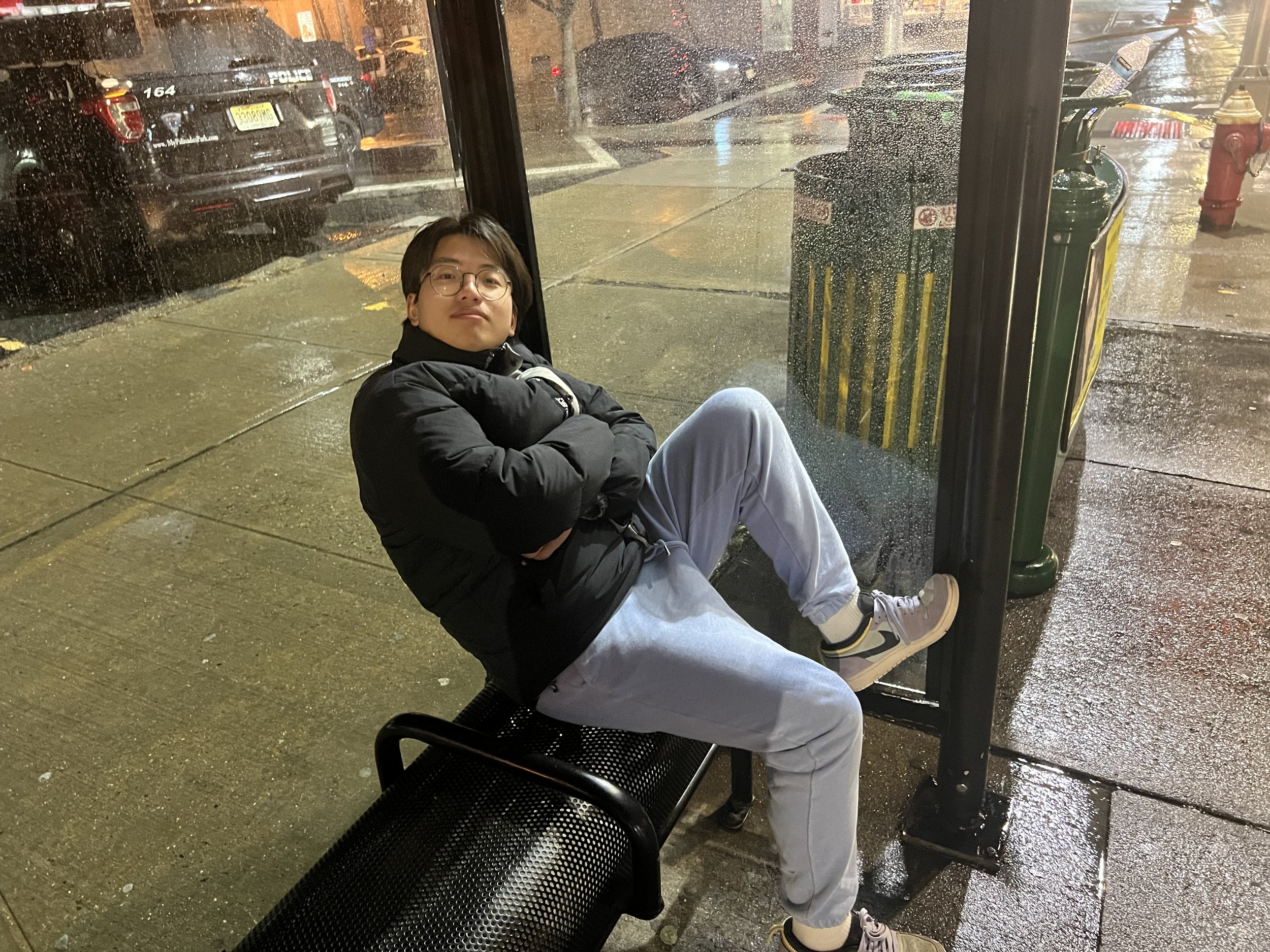 A young man sitting on a bench at a bus stop on a rainy night, looking at the camera with his arms crossed. Rain is visible on the street and window glass, with cars parked along the street and a fire hydrant nearby.