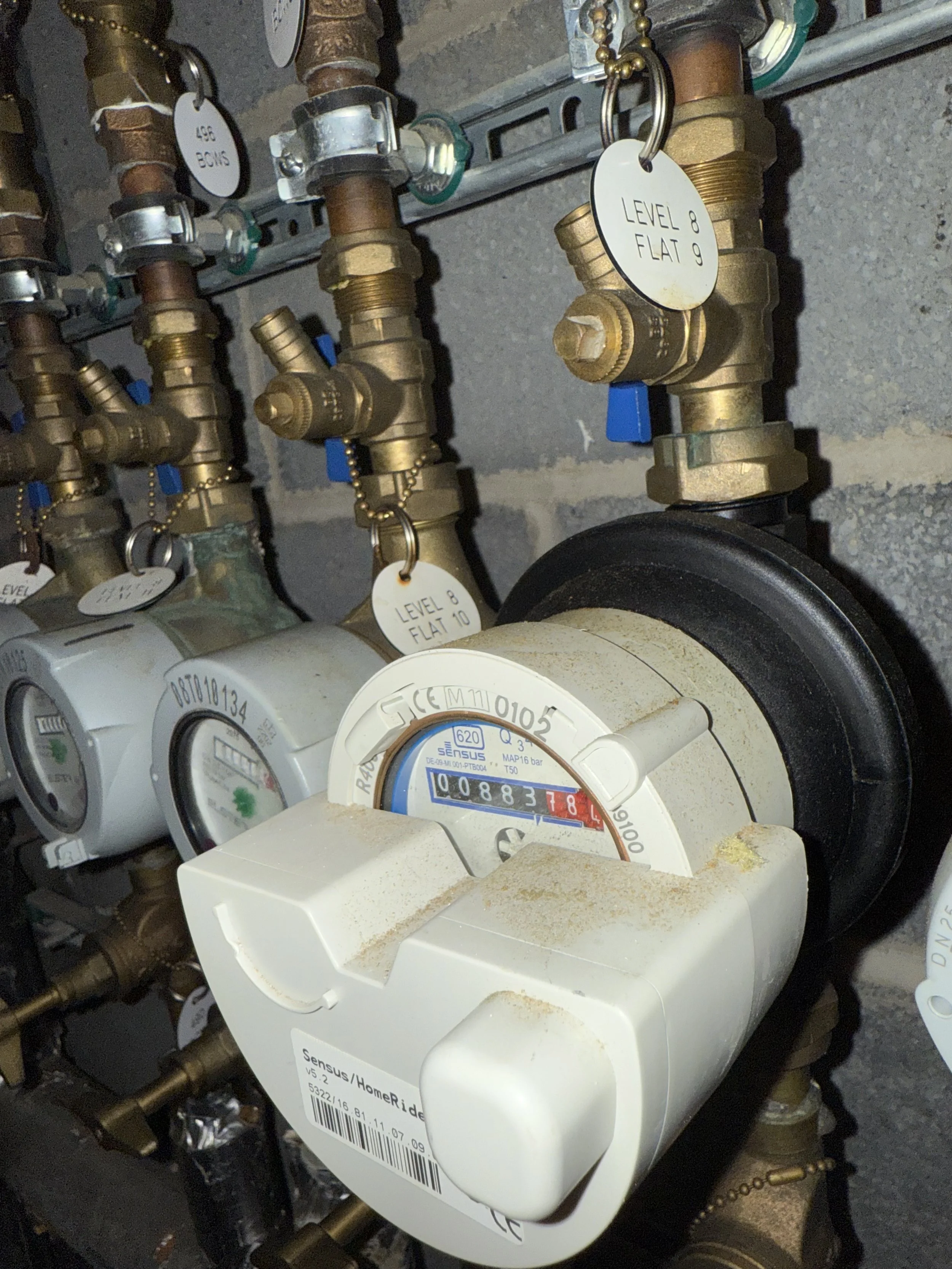 A collection of water meters and plumbing valves with labels indicating levels and flat settings, installed on a wall.