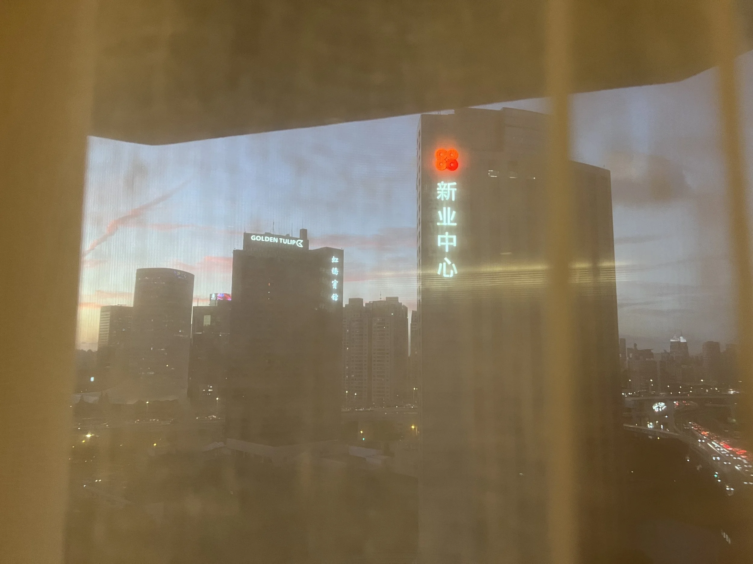 City skyline at dusk with high-rise buildings, illuminated signs in Chinese characters and English, including 'GOLDEN TULIP', under a cloudy sky.