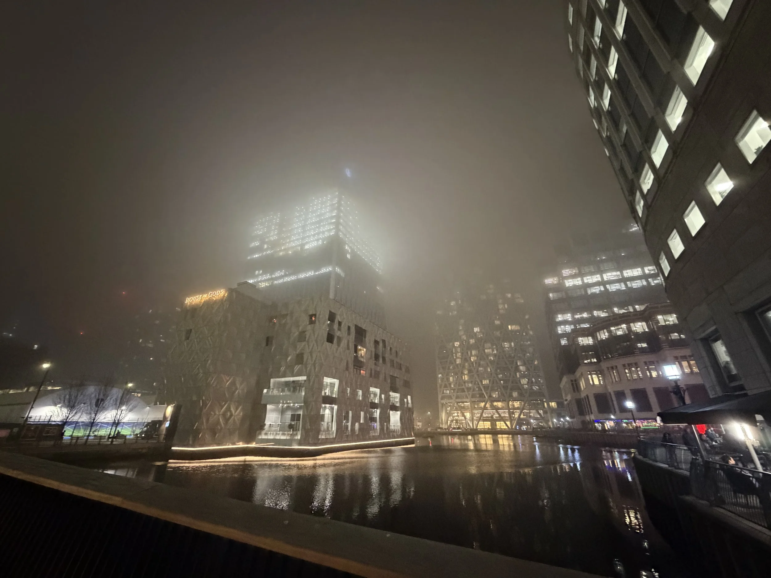 Nighttime cityscape with tall buildings, some with illuminated windows, on a foggy or rainy night. Reflections on wet surfaces and a visible sign reading 'HOUSE OF GOD' on one of the buildings.