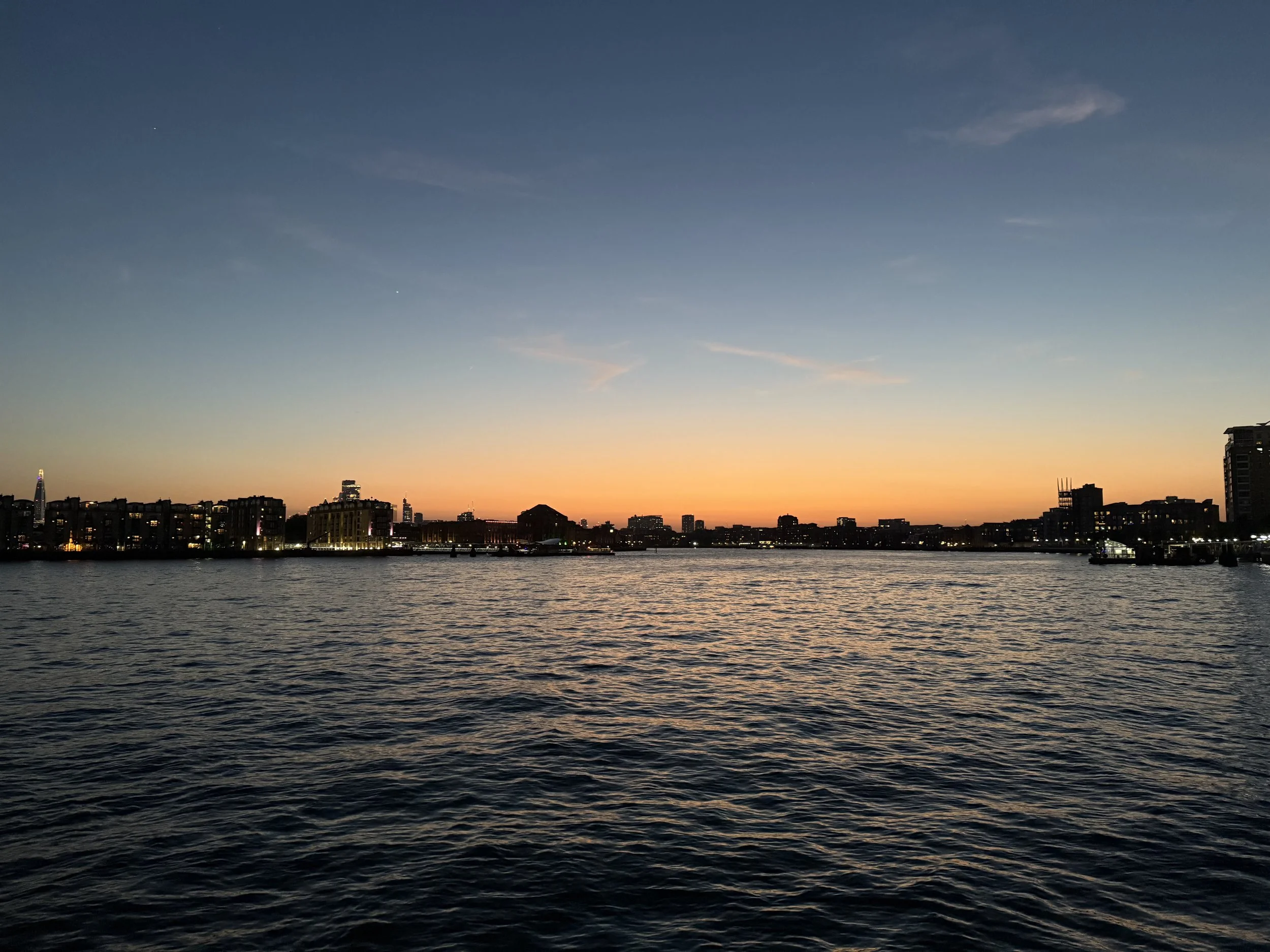 City skyline at sunset with buildings along a body of water, clear sky with a few clouds, and warm colors near the horizon.