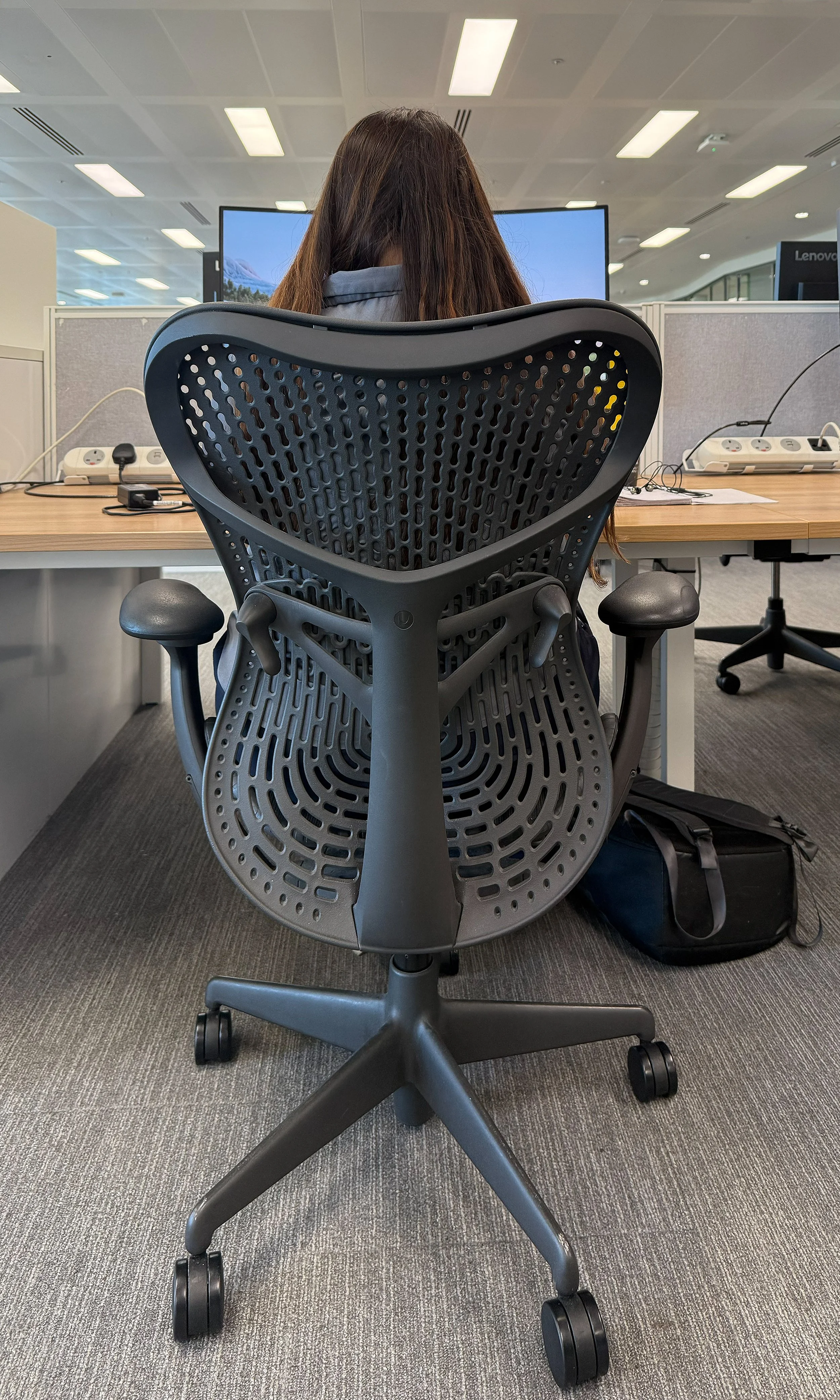 Woman with long brown hair sitting in a black ergonomic office chair at a desk, facing a large monitor in an open office with overhead fluorescent lighting.