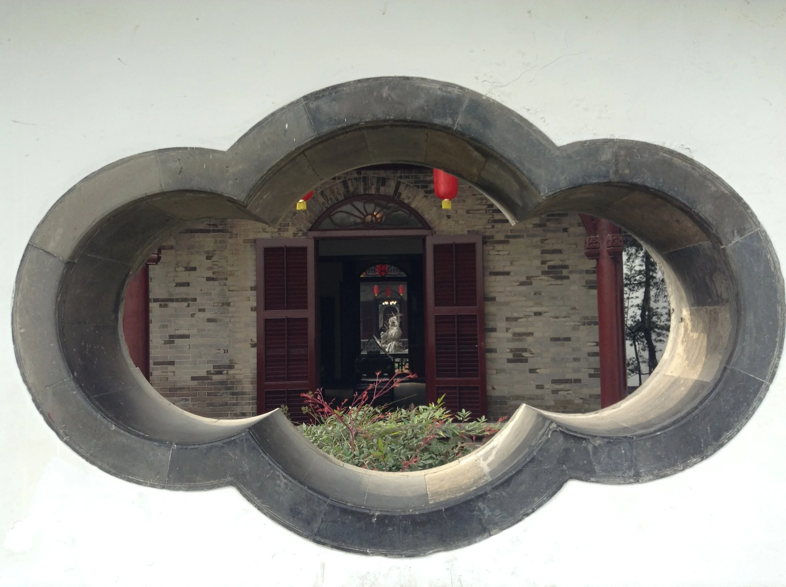 View through a circular opening in a louvered wall showing a doorway with an arched window, brick walls, red lanterns hanging from the ceiling, and plants in front.