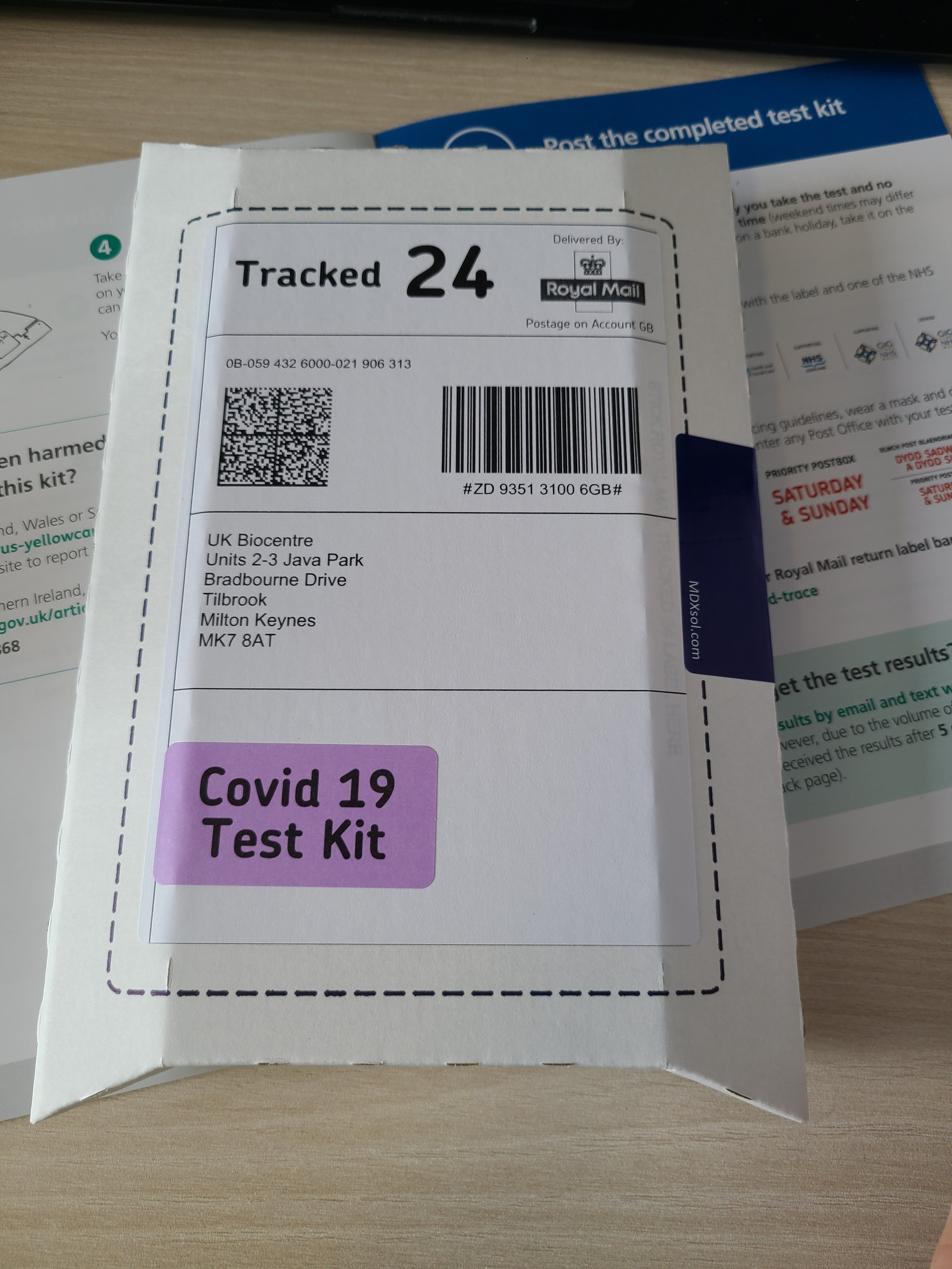 A postal package with a tracking label attached, indicating it has been delivered by Royal Mail, addressed to UK Biocentre in Milton Keynes, with a purple Covid-19 test kit label on the front.