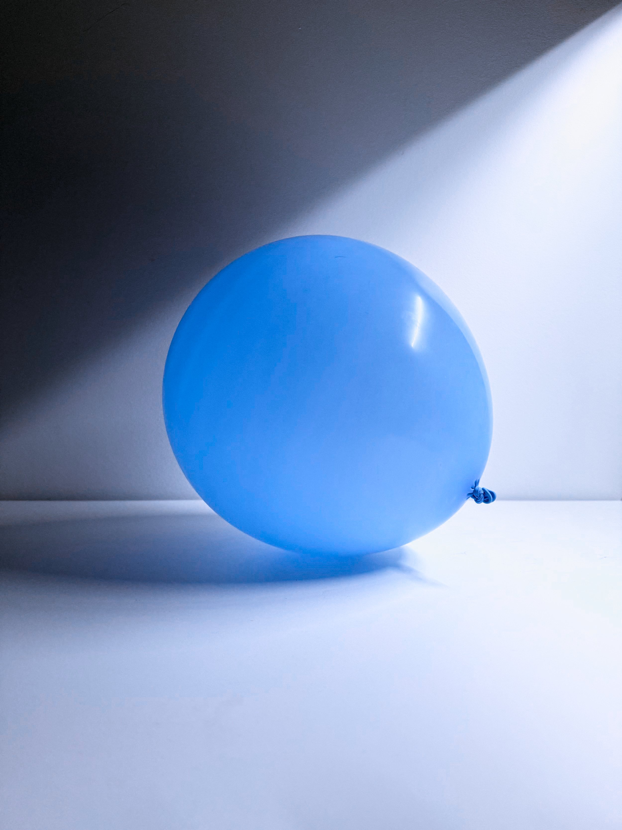A blue balloon resting on a white surface with a shadow cast behind it, illuminated by bright light from the right side.