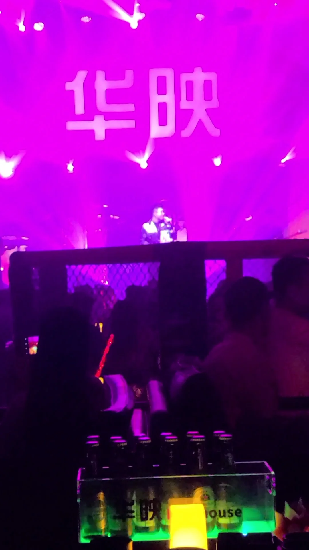 People sitting at tables inside a nightclub with purple lighting. A DJ is performing on stage with a large pink neon sign featuring Chinese characters behind him. Bottled drinks are visible on the table in the foreground, illuminated by colorful ligh