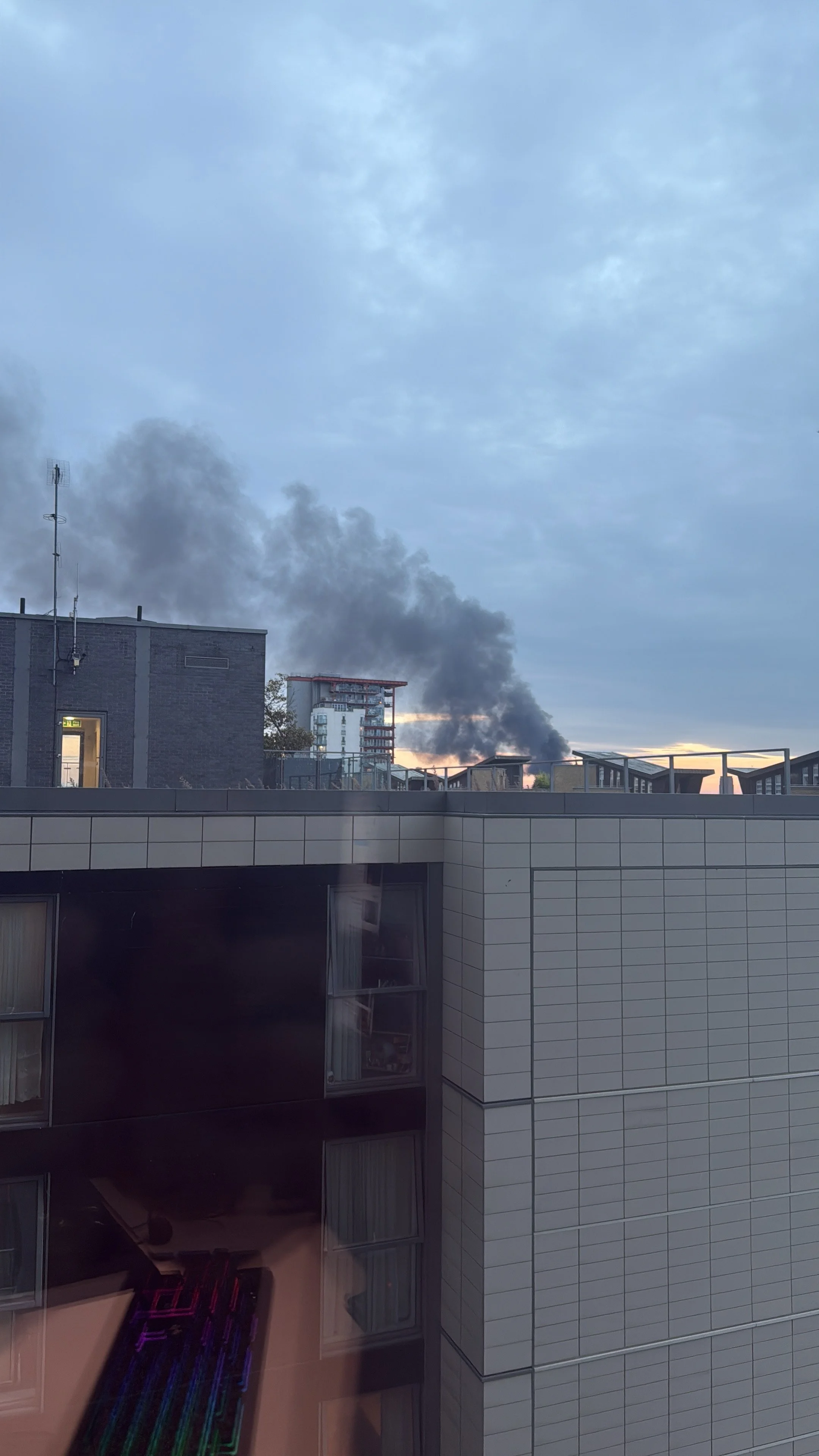 Smoke rising from a building in an urban area during sunset, with neighboring buildings and a partly cloudy sky.
