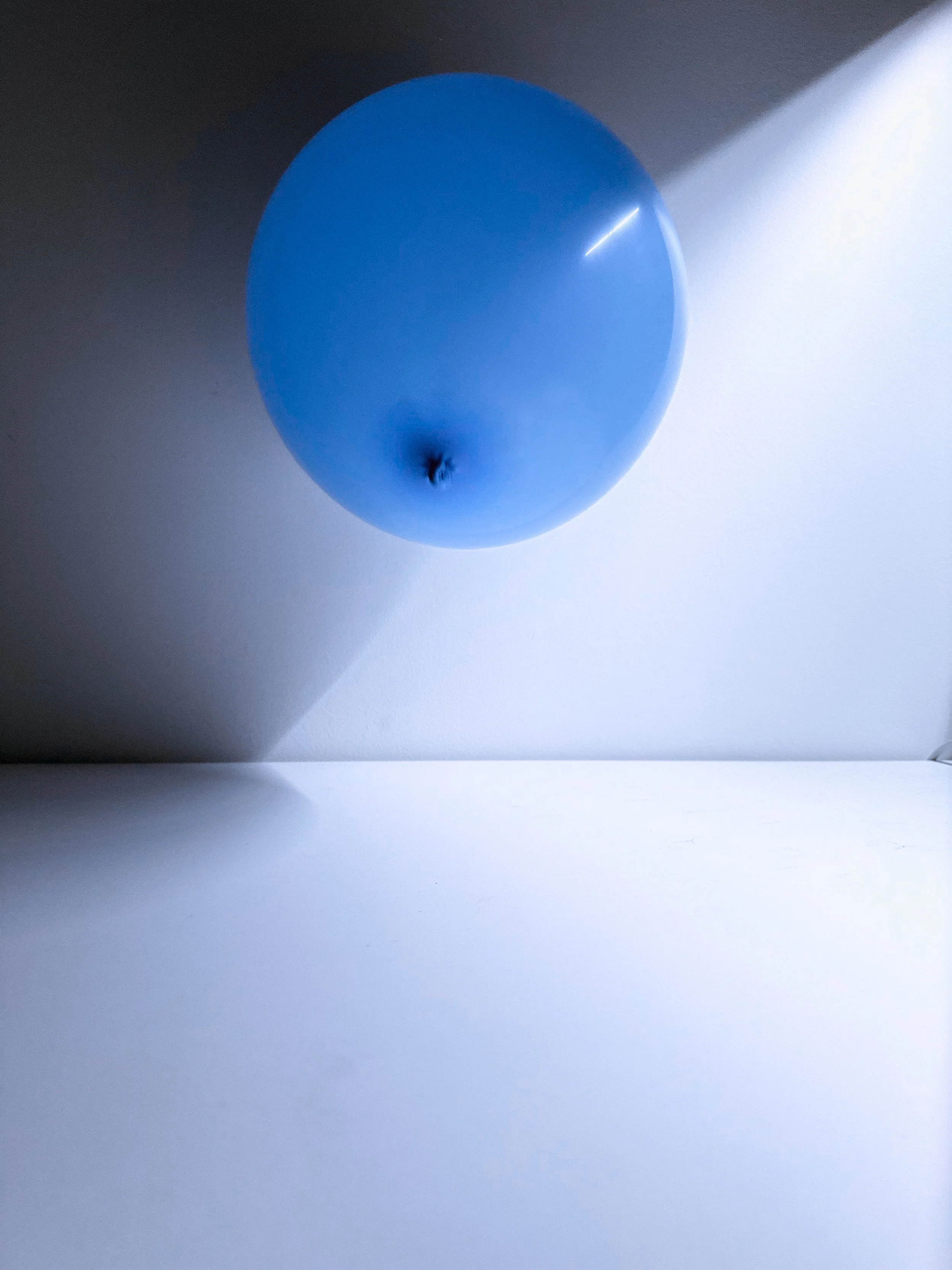 A blue balloon illuminated by a narrow beam of light, casting shadows on a white surface and the wall behind it.