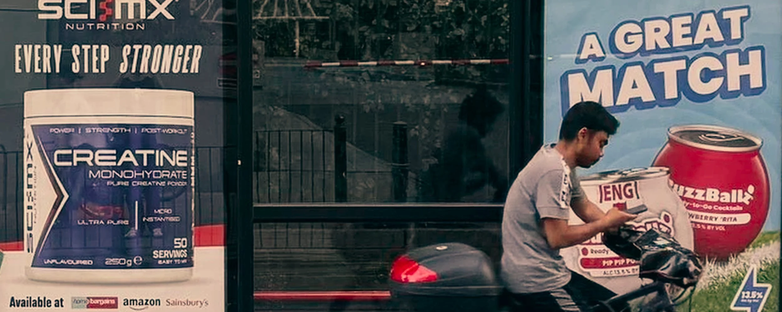 A man on a bicycle looking at his phone next to large advertisements for health supplements and snacks on a store window.