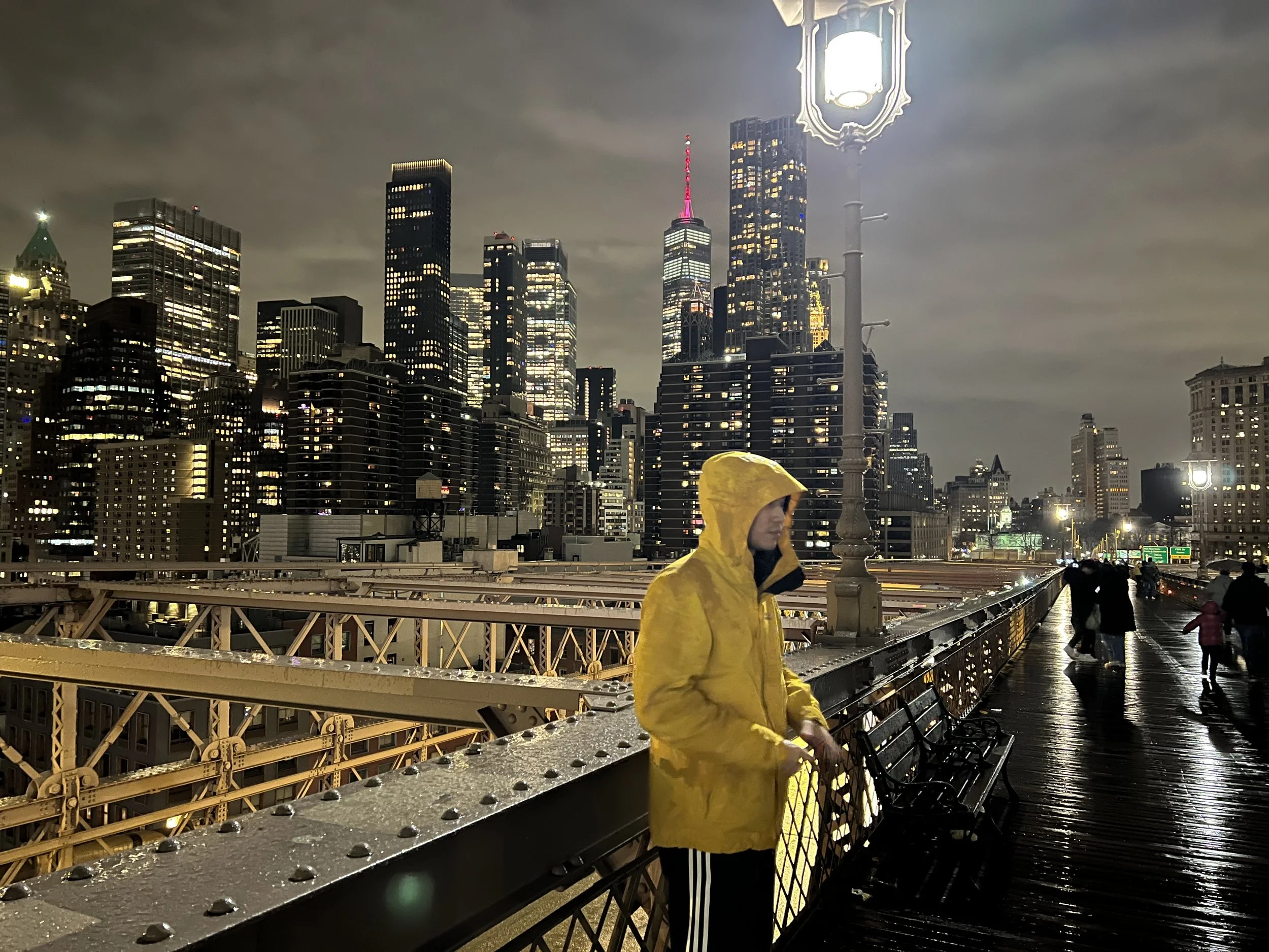 Nighttime city skyline with tall lit-up buildings, including the Empire State Building partly lit in pink, seen from a bridge with benches and street lamps. A person in a yellow raincoat is walking on the bridge.