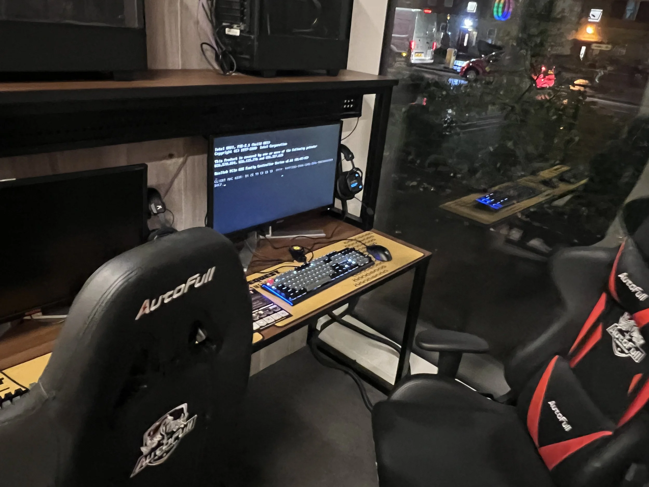A computer workstation setup near a window with a view of a parking lot and street outside. The setup includes a monitor displaying error text, a keyboard, a mouse, and a headset on a wooden desk. There is a racing car-style seat in the foreground an