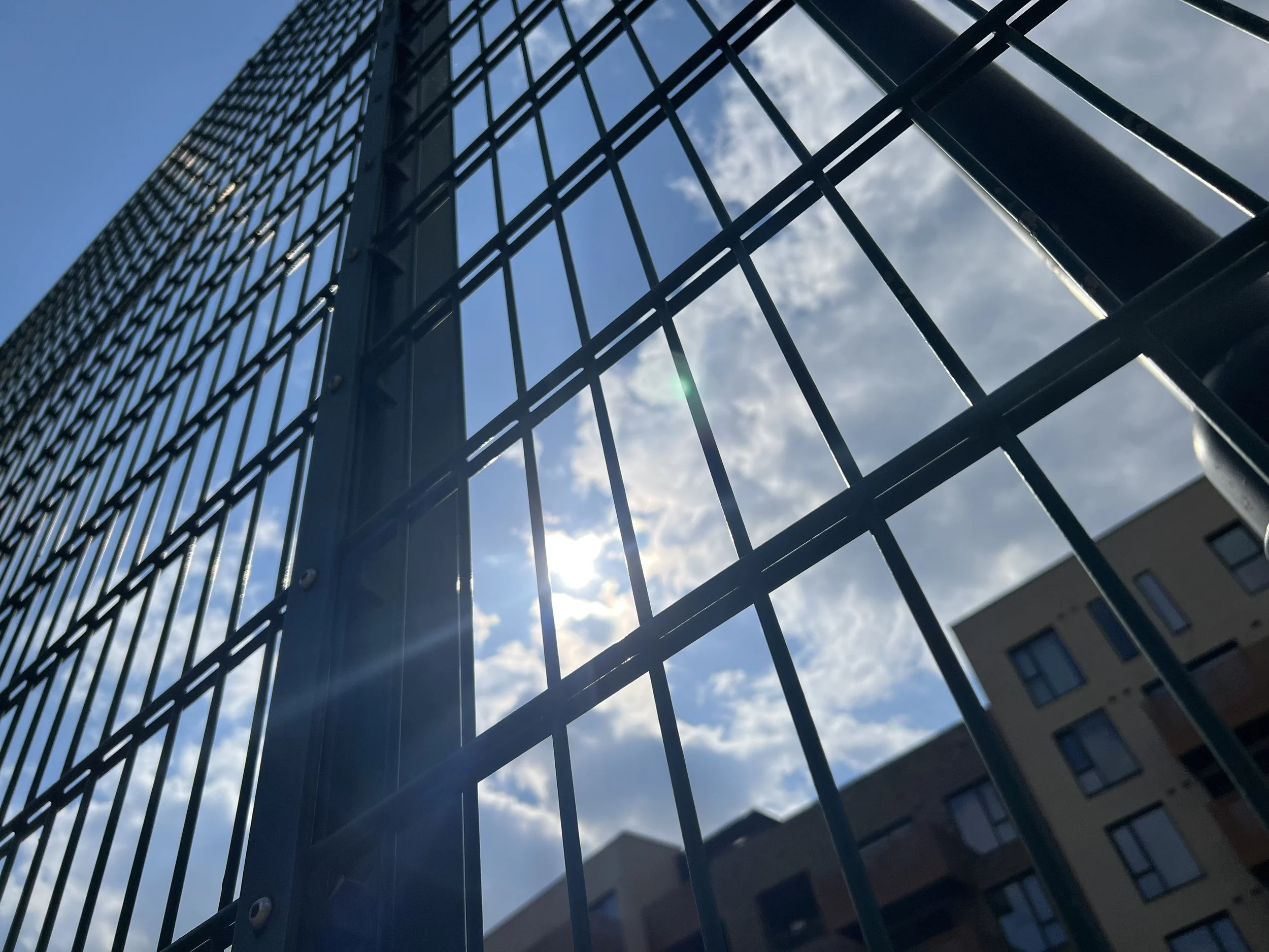 Looking up at a modern glass building reflecting the sky and clouds, with a metal fence in the foreground.