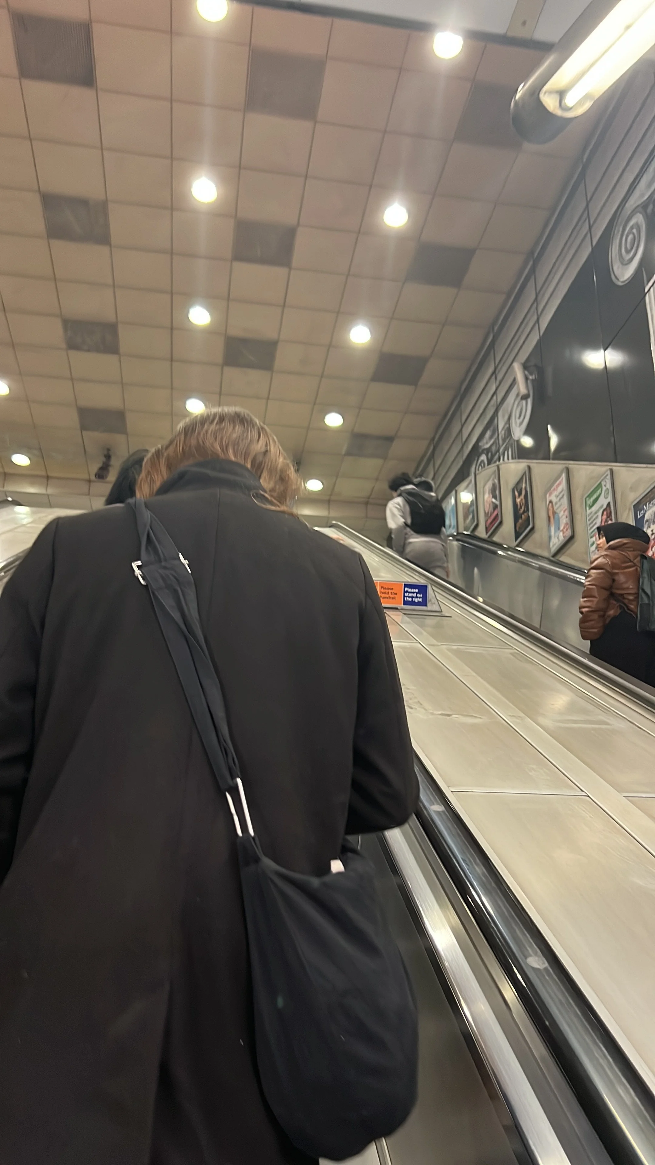 People riding an escalator in a subway station with ceiling lights and advertisements.