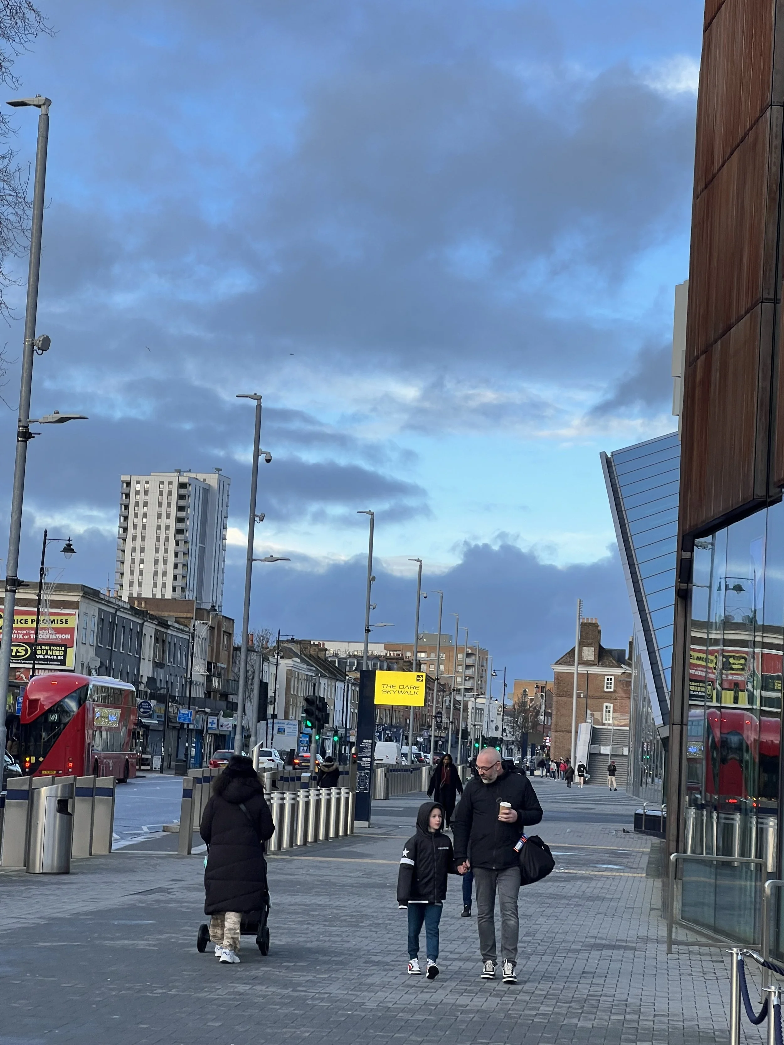 People walking in a busy city street with tall buildings, streetlights, and a red double-decker bus, under a cloudy sky.