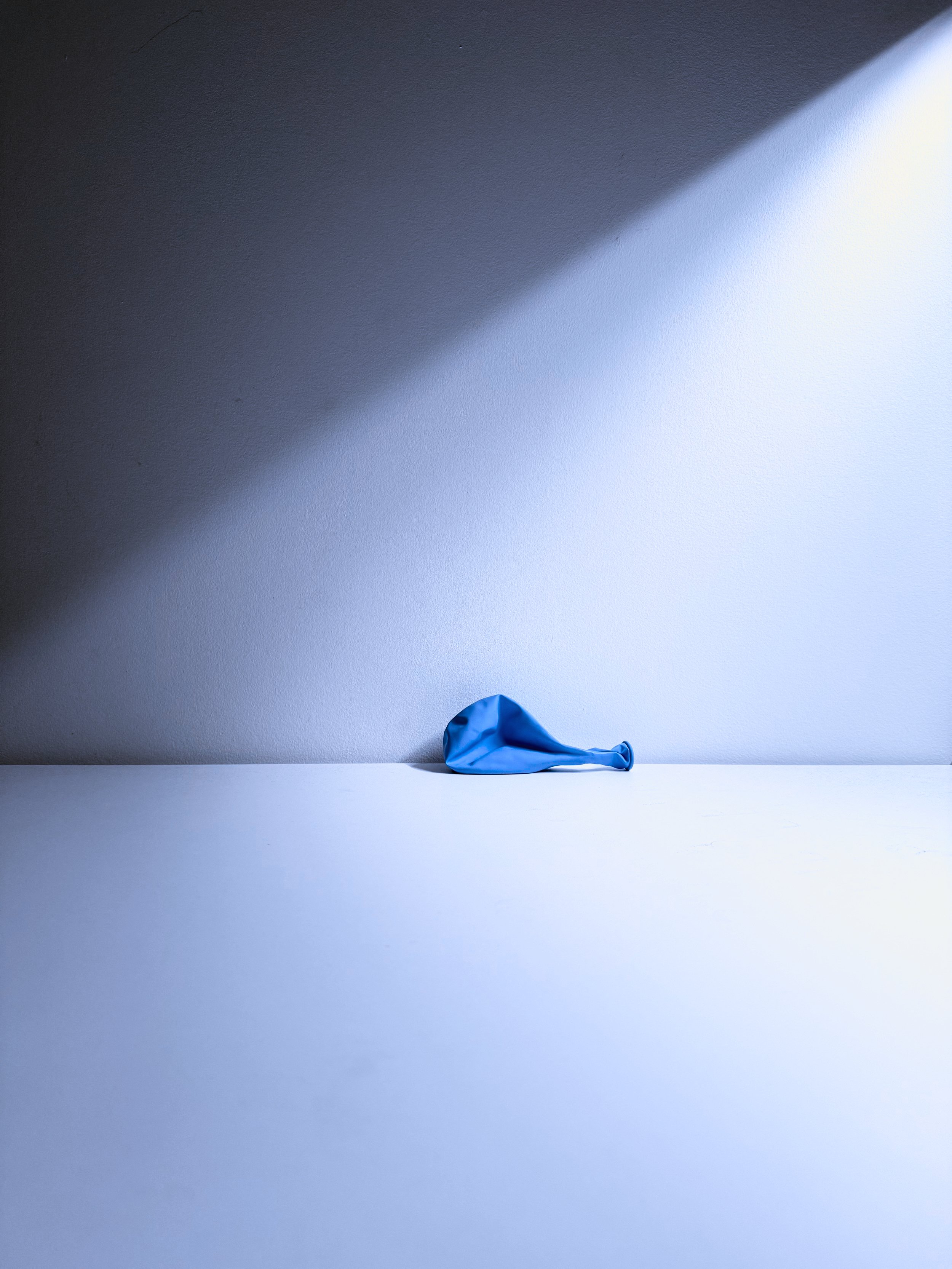 A deflated blue balloon resting on a white surface, casting a shadow on a light blue wall.