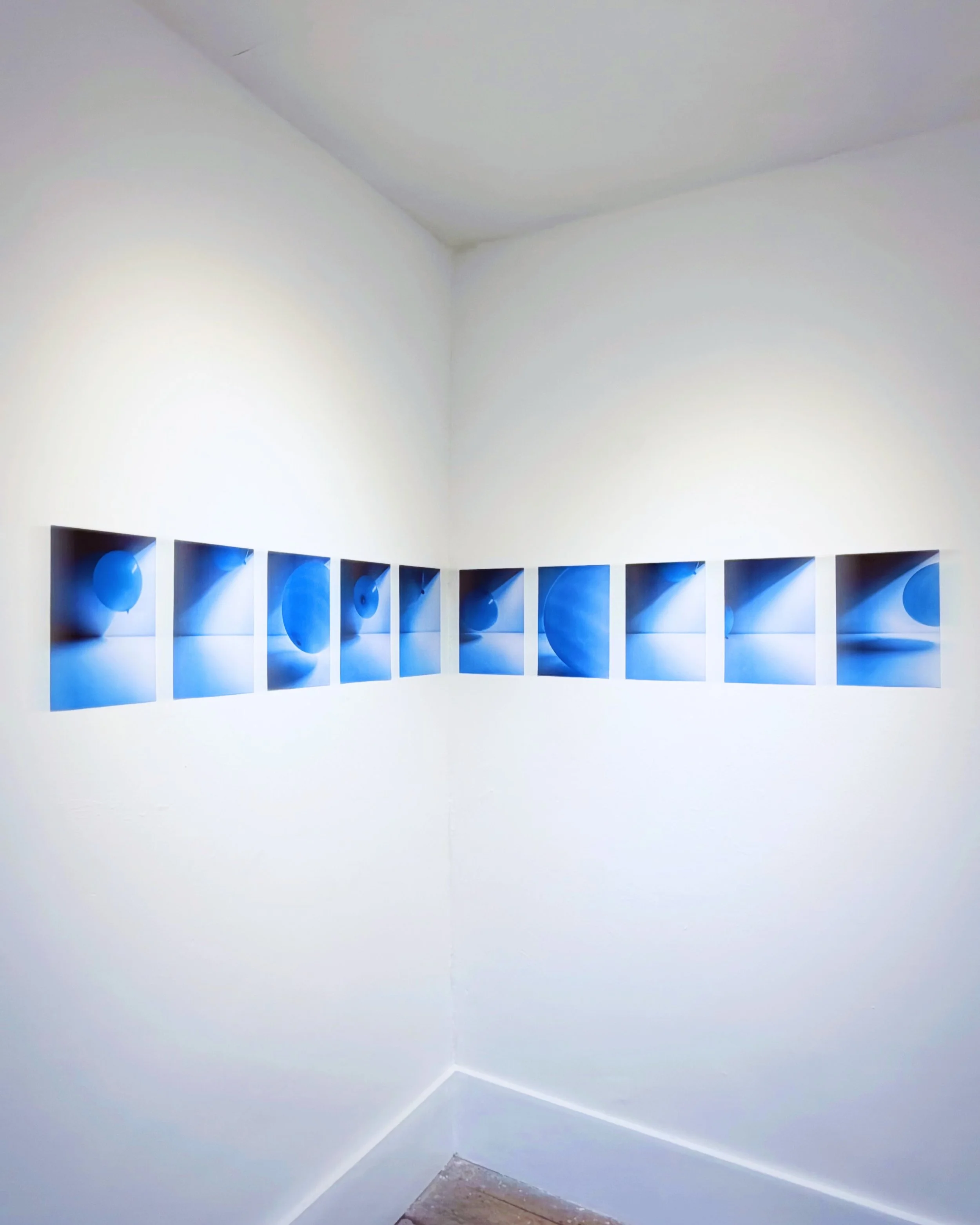Empty corner of a room with white walls, ceiling, and baseboards, with a row of blue abstract paintings on the lower part of the wall.