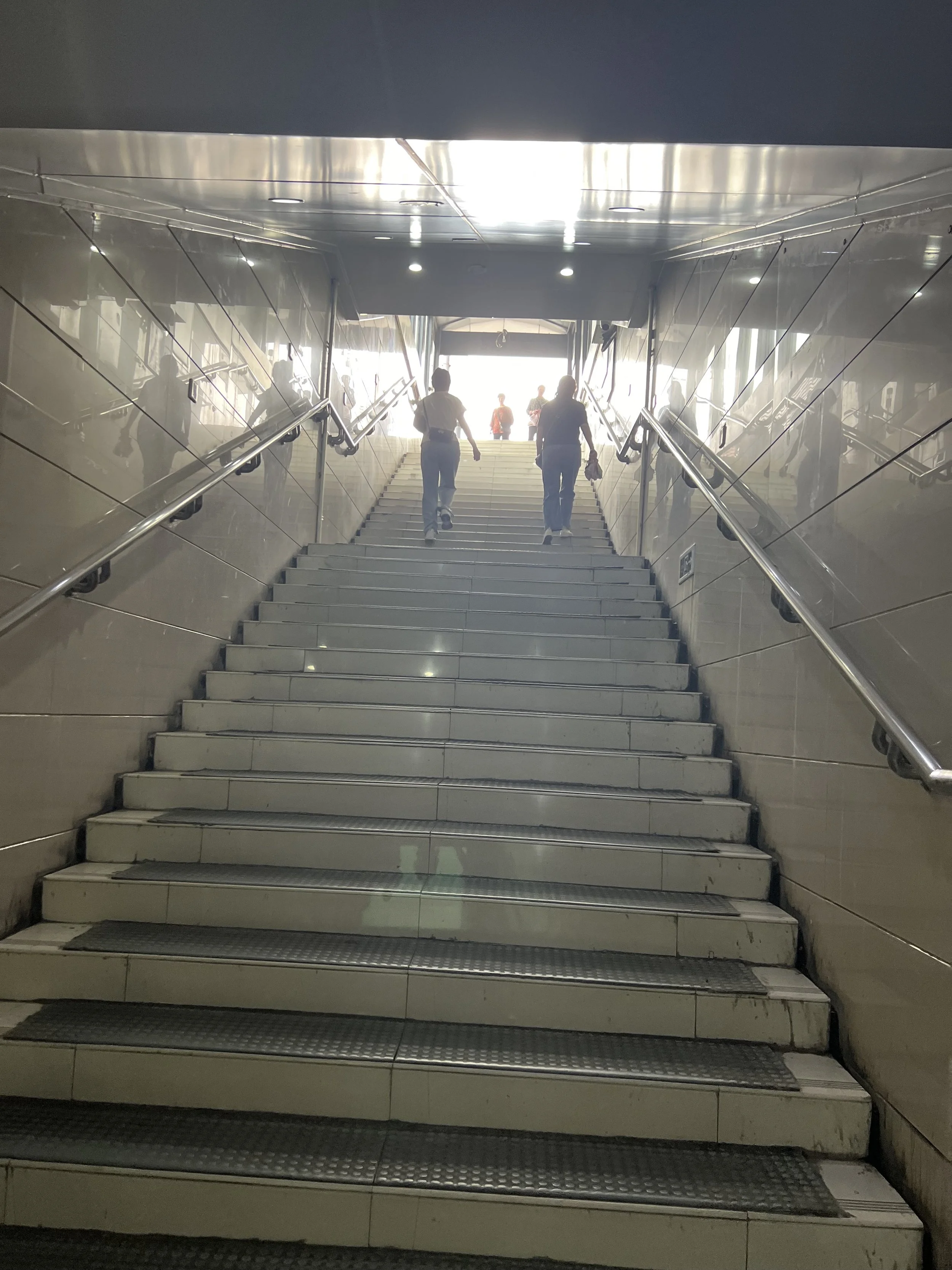 People ascending a staircase in an indoor setting with reflective, polished wall surfaces and bright light at the top of the stairs.
