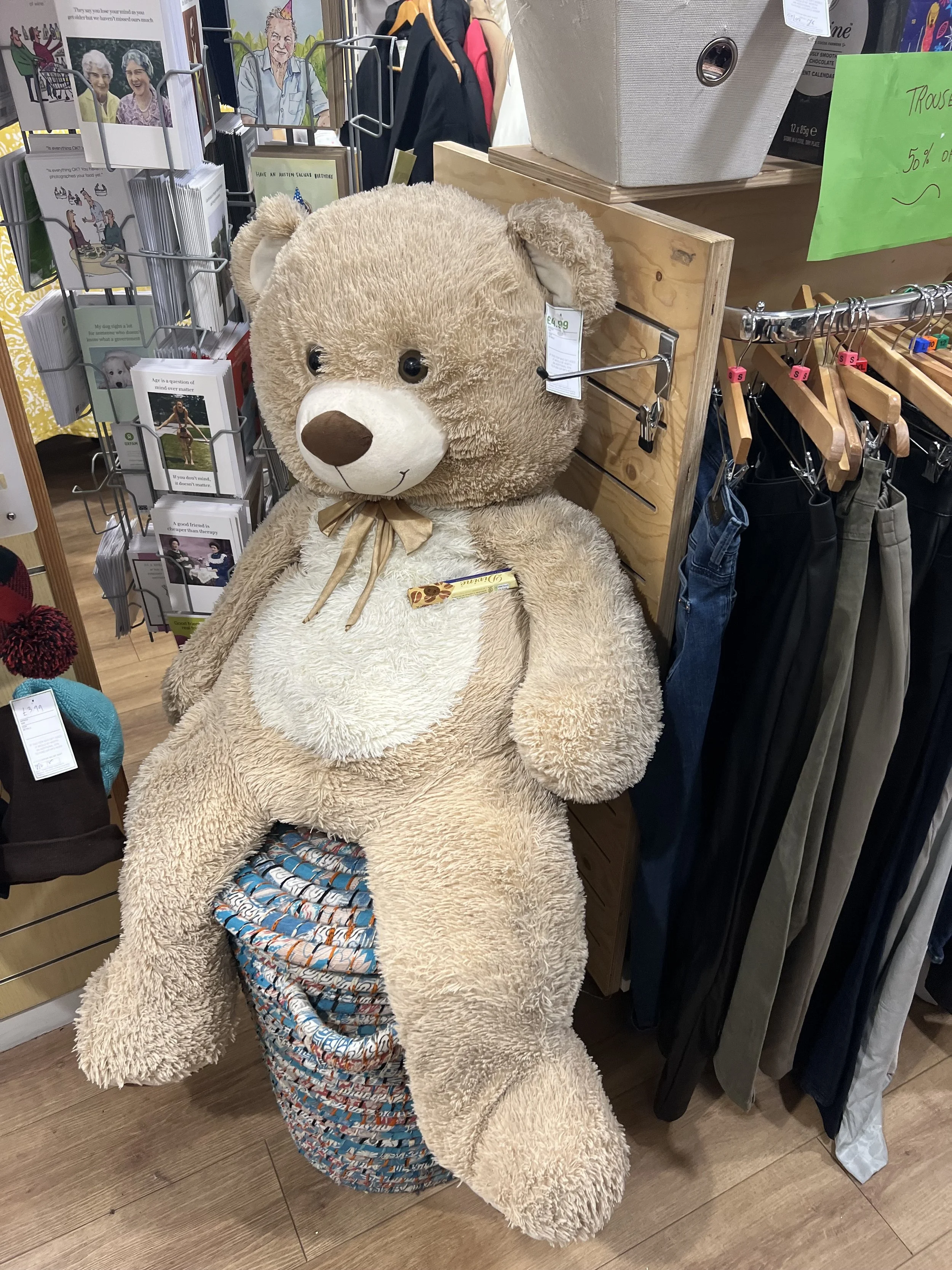 A large teddy bear sitting on a chair with a colorful blanket pattern on its lap, surrounded by clothes and greeting cards in a store.