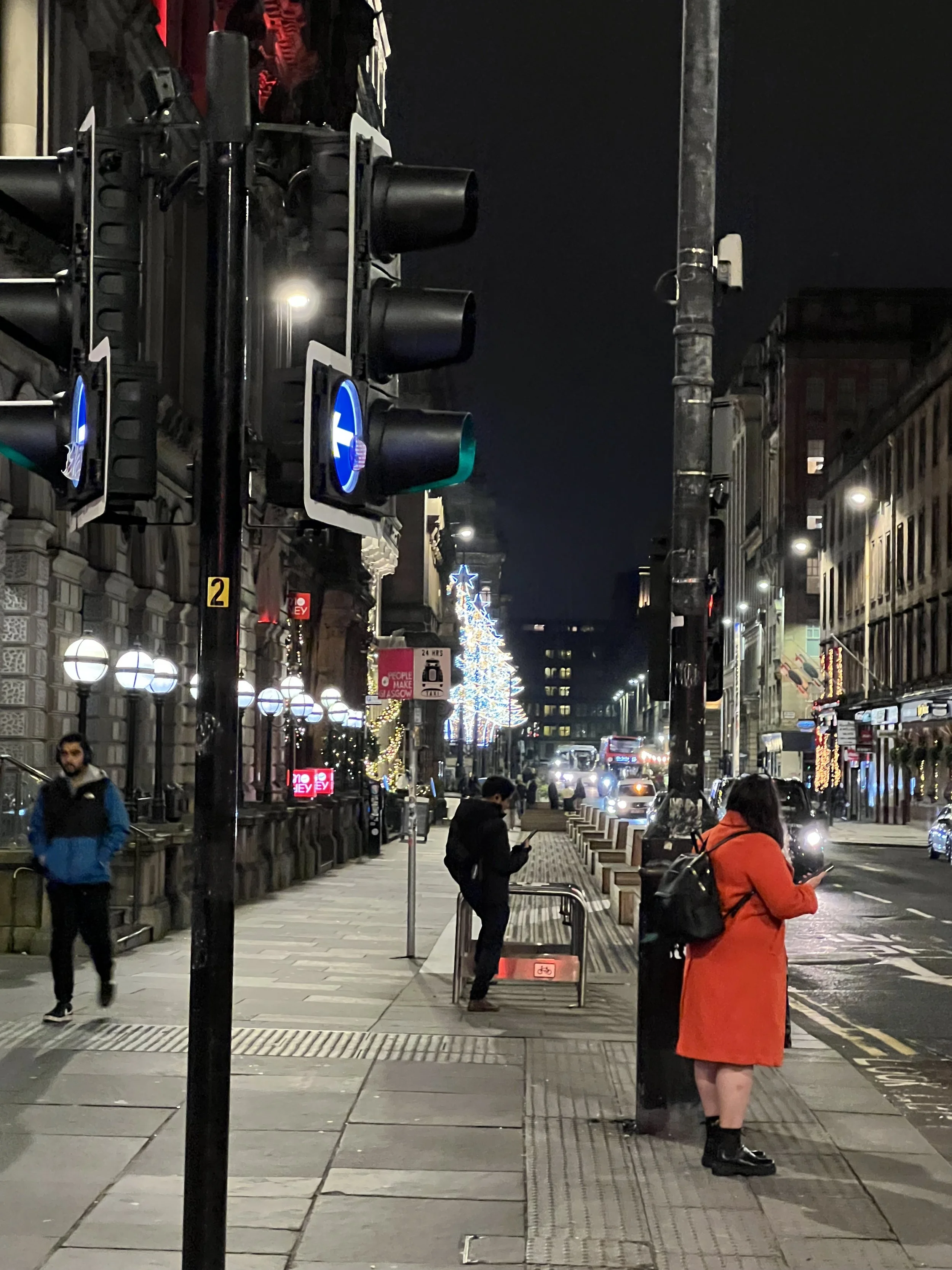 Nighttime street scene with pedestrians and traffic lights, decorated with holiday lights and a Christmas tree in the distance, in an urban area.