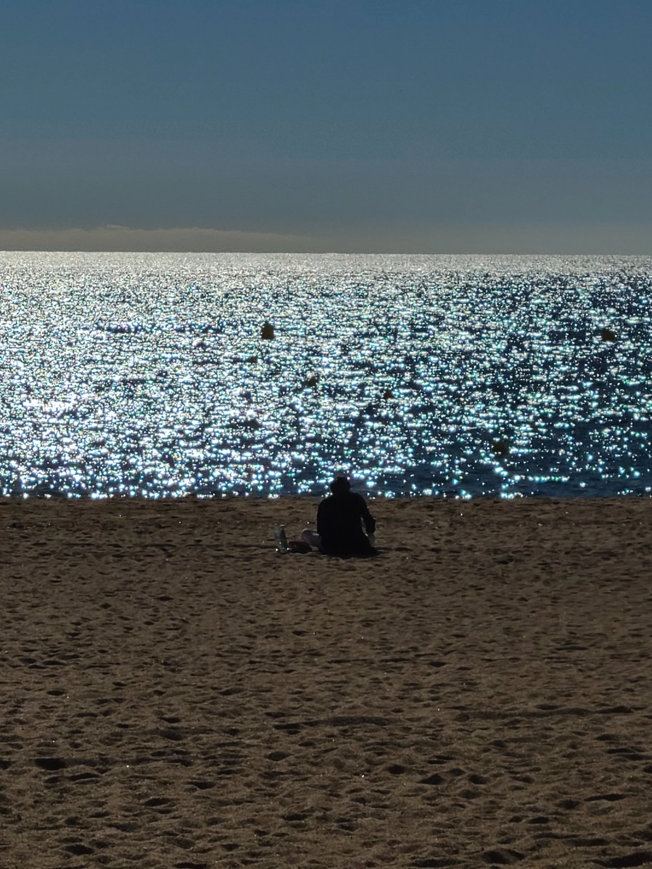 A person sitting on a sandy beach facing the ocean with a glittering water surface and a dark sky.