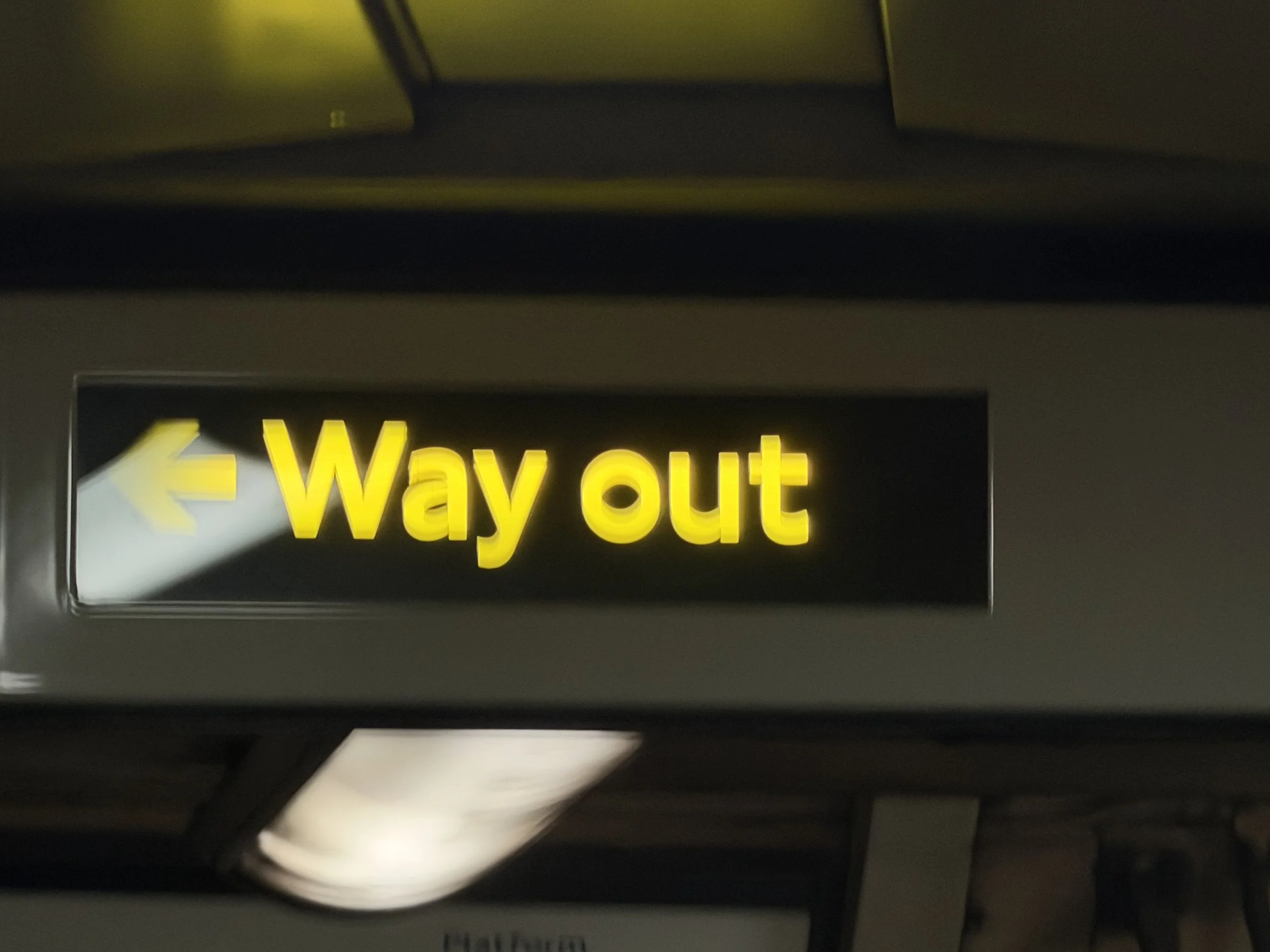 Sign with yellow text on a black background indicating the way out, with a yellow arrow pointing to the left.